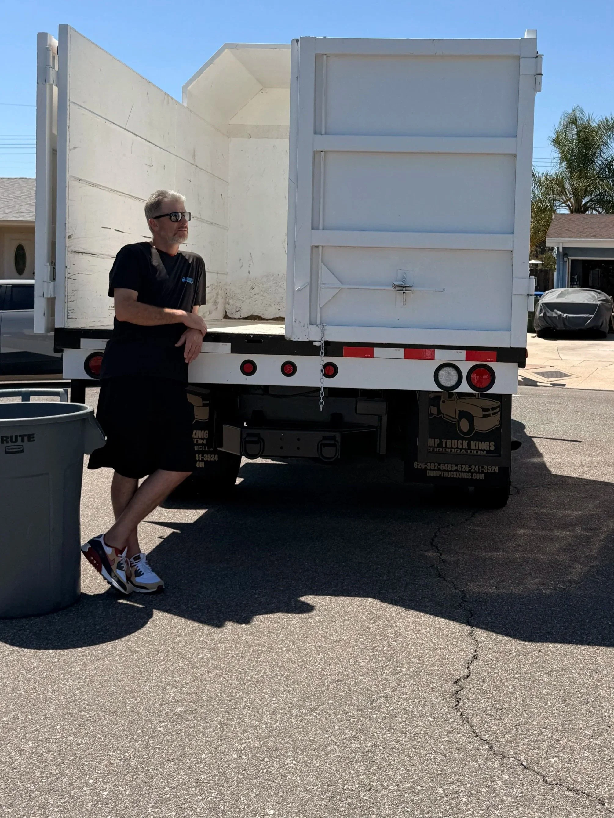 A man with gray hair, a beard, wearing sunglasses, a black t-shirt, shorts, and sneakers, leaning against the side of a white truck with an open back, in a suburban neighborhood during the daytime.