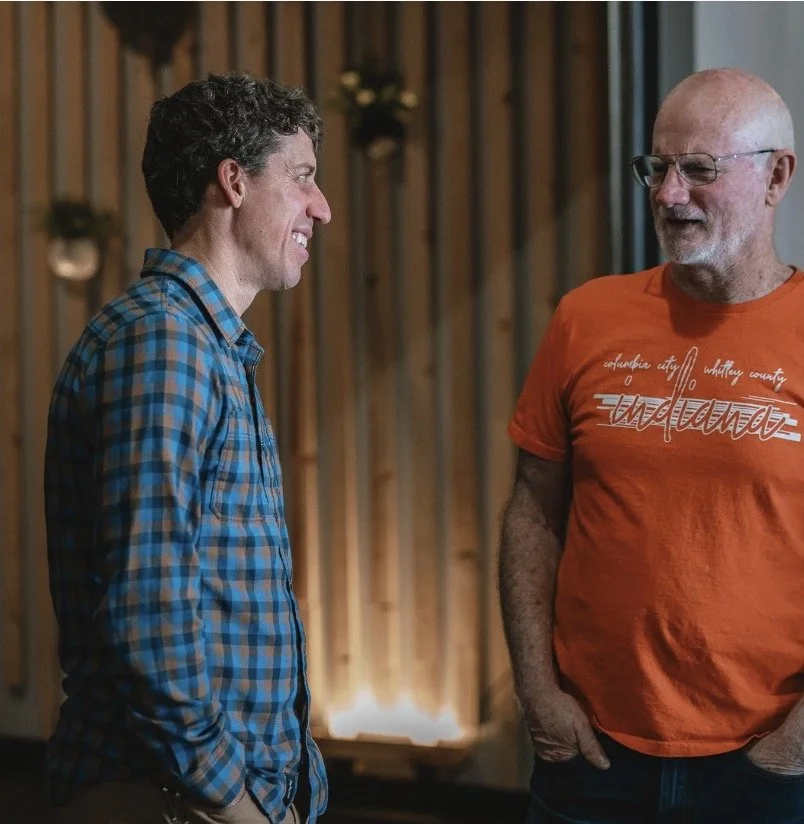 Two men smiling and talking indoors. One man has curly hair and is wearing a blue plaid shirt. The other is bald with glasses and wearing an orange t-shirt that says 'Florida'.