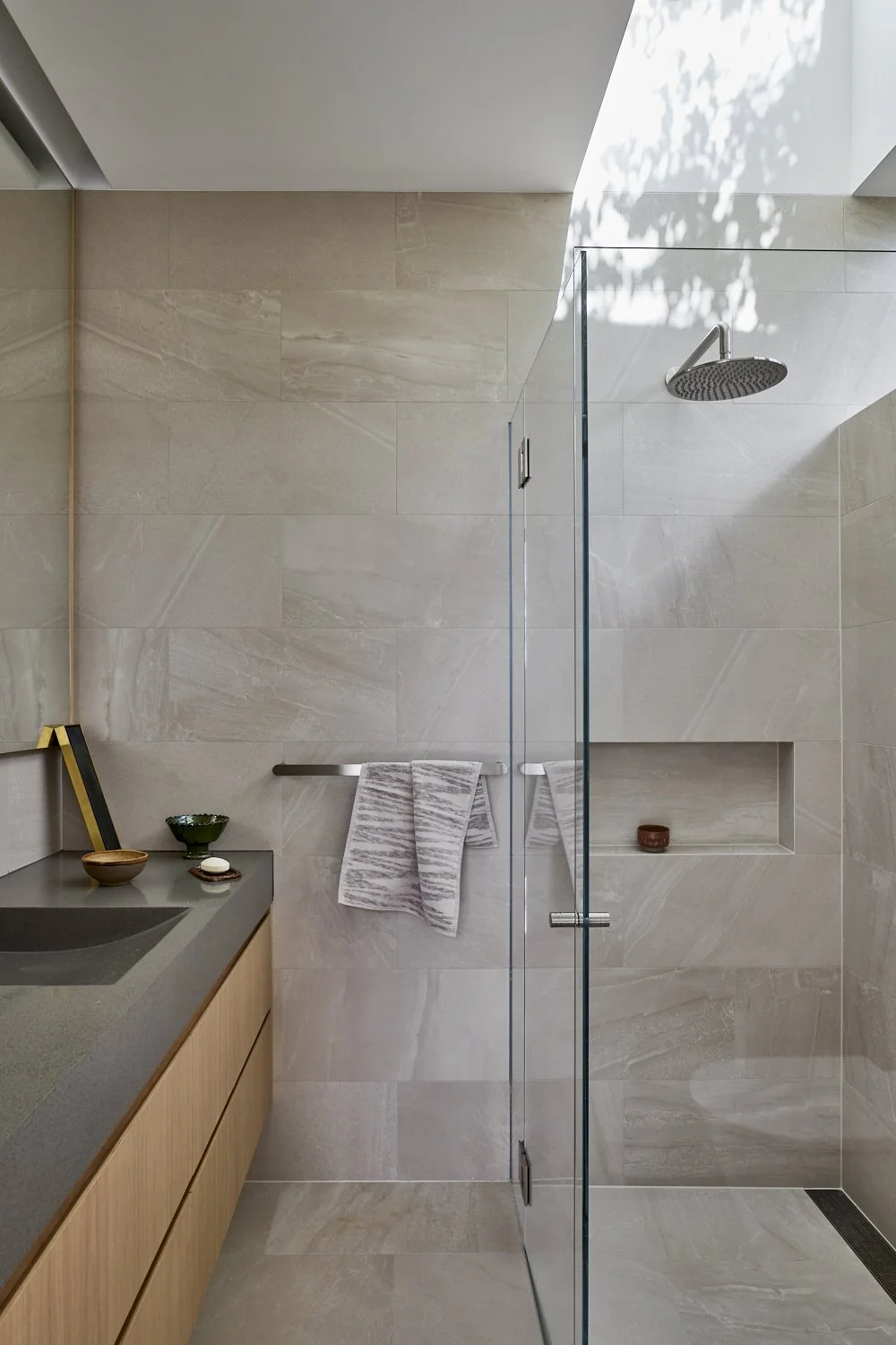 Modern bathroom with beige stone walls and shower area featuring a rain showerhead and a built-in shelf, with towels hanging on a wall hook and a wooden vanity with bowls and framed artwork.