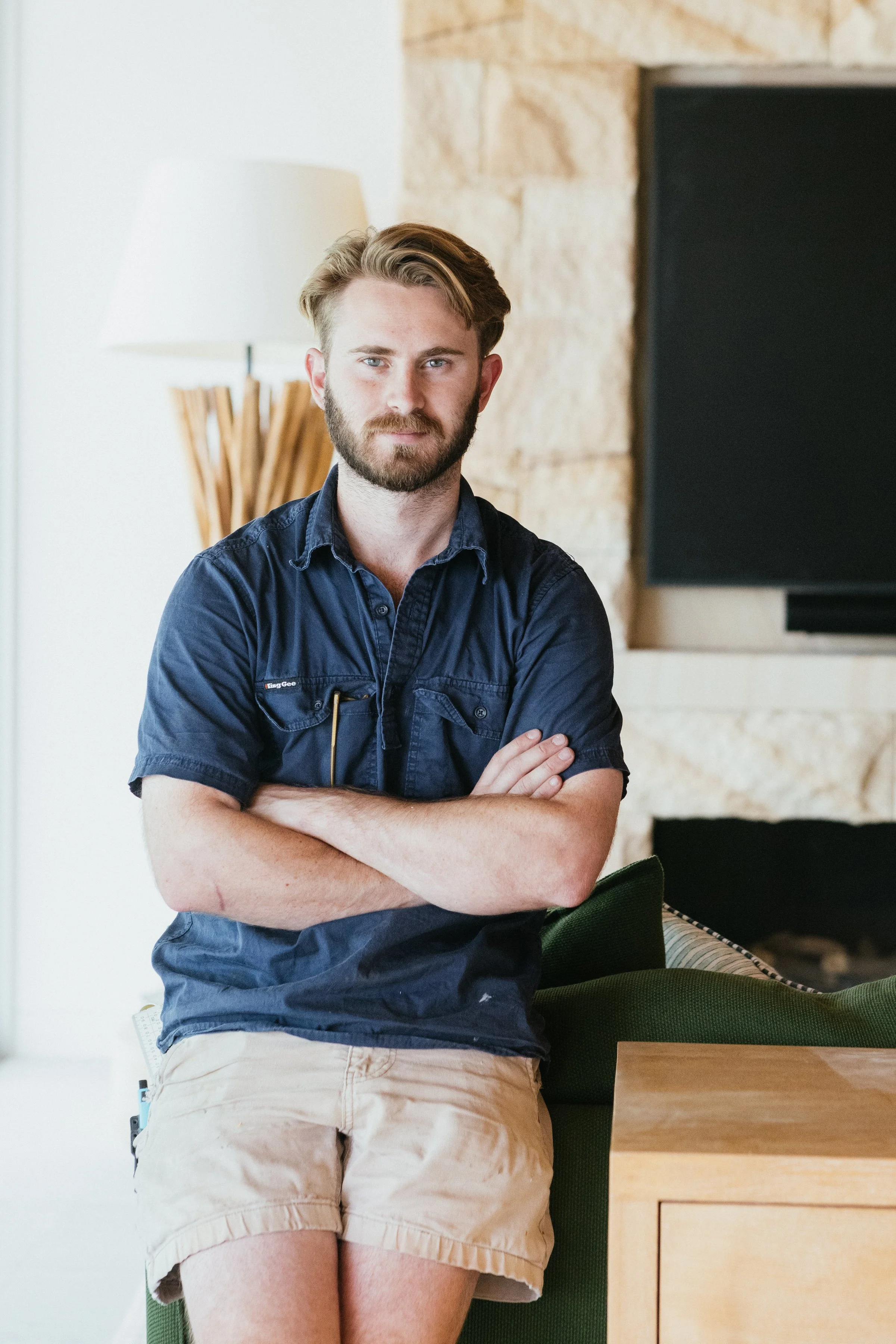 A young man with a beard and blond hair, wearing a navy blue shirt and tan shorts, standing with arms crossed in a contemporary living room.