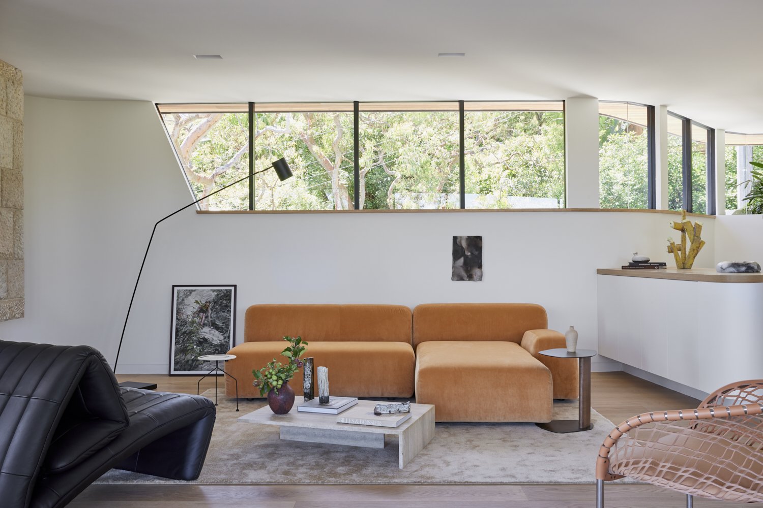 Living room with orange sofa, black leather chair, beige coffee table with vases, art and greenery decor, large windows with trees outside, modern minimalist design