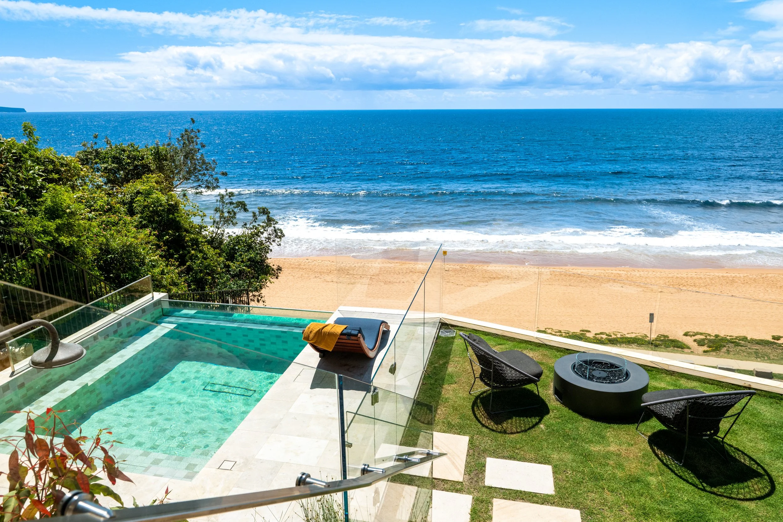 View of a coastal scene with a swimming pool in the foreground, lush greenery, sandy beach, and the ocean with blue water and a partly cloudy sky in the background.