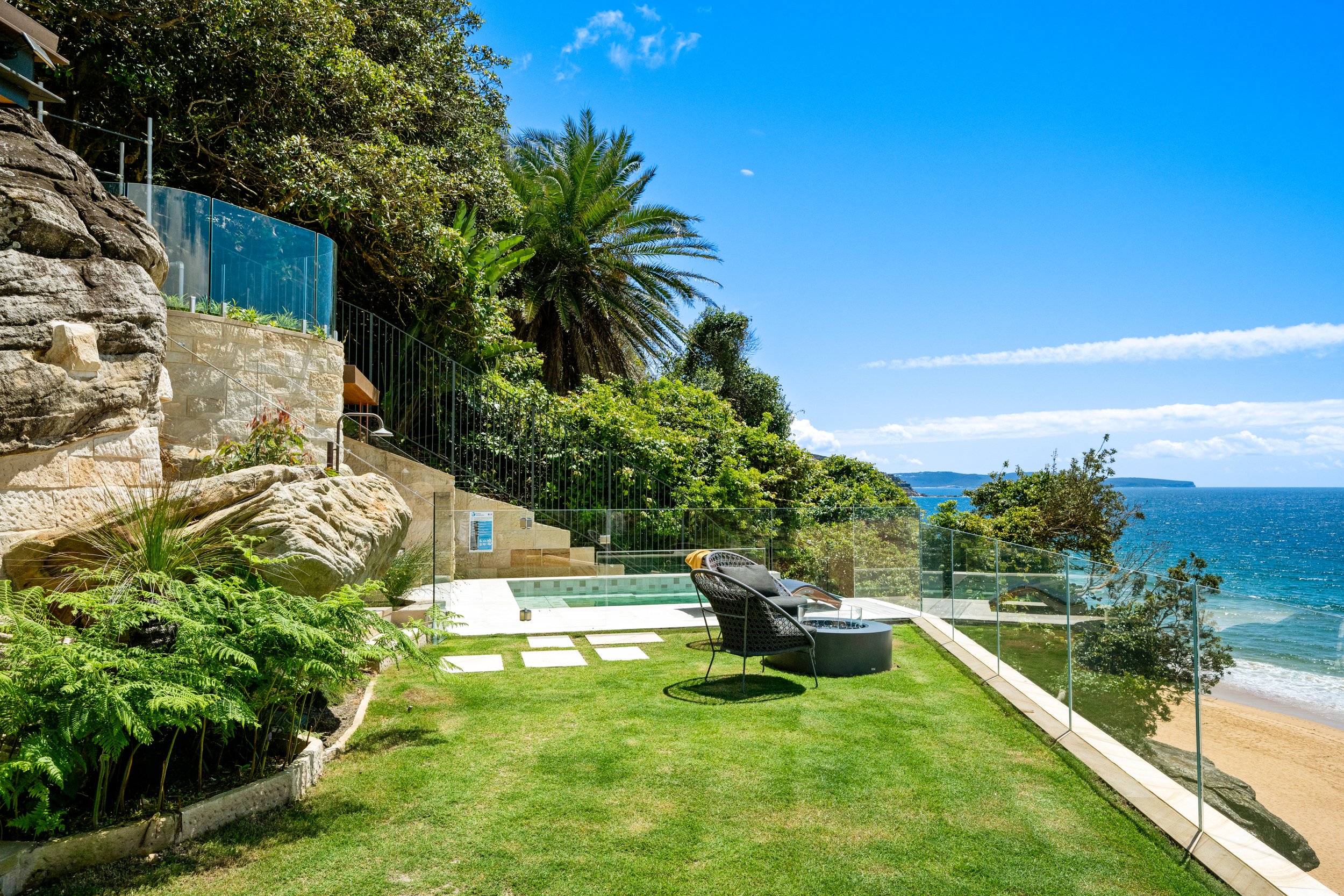 Poolside backyard with glass fencing overlooking the ocean, surrounded by lush green plants, trees, and seating area on a sunny day.