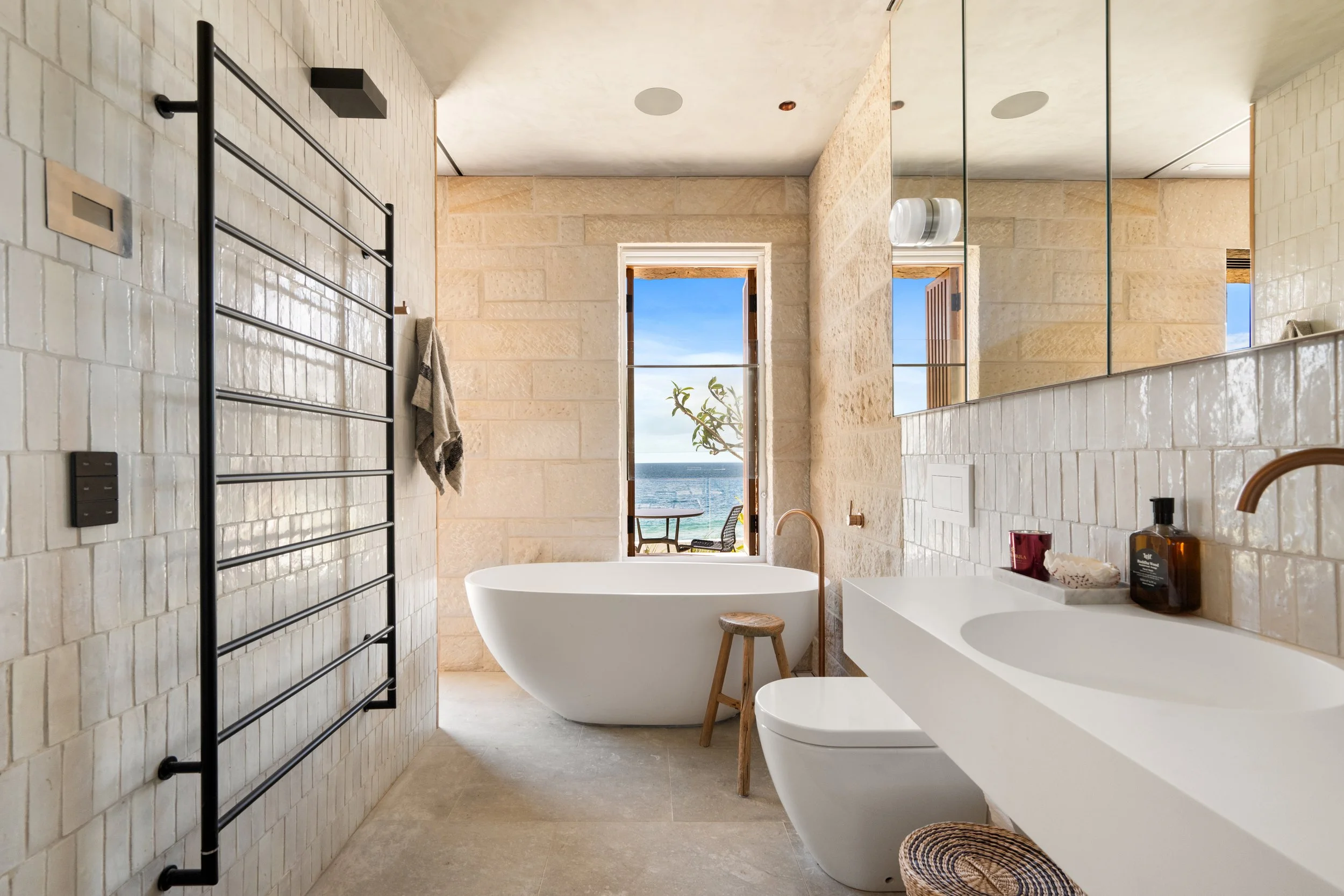 Modern bathroom with a freestanding bathtub, dual sinks, large mirror, and a window with ocean view, beige brick walls, and natural decor.
