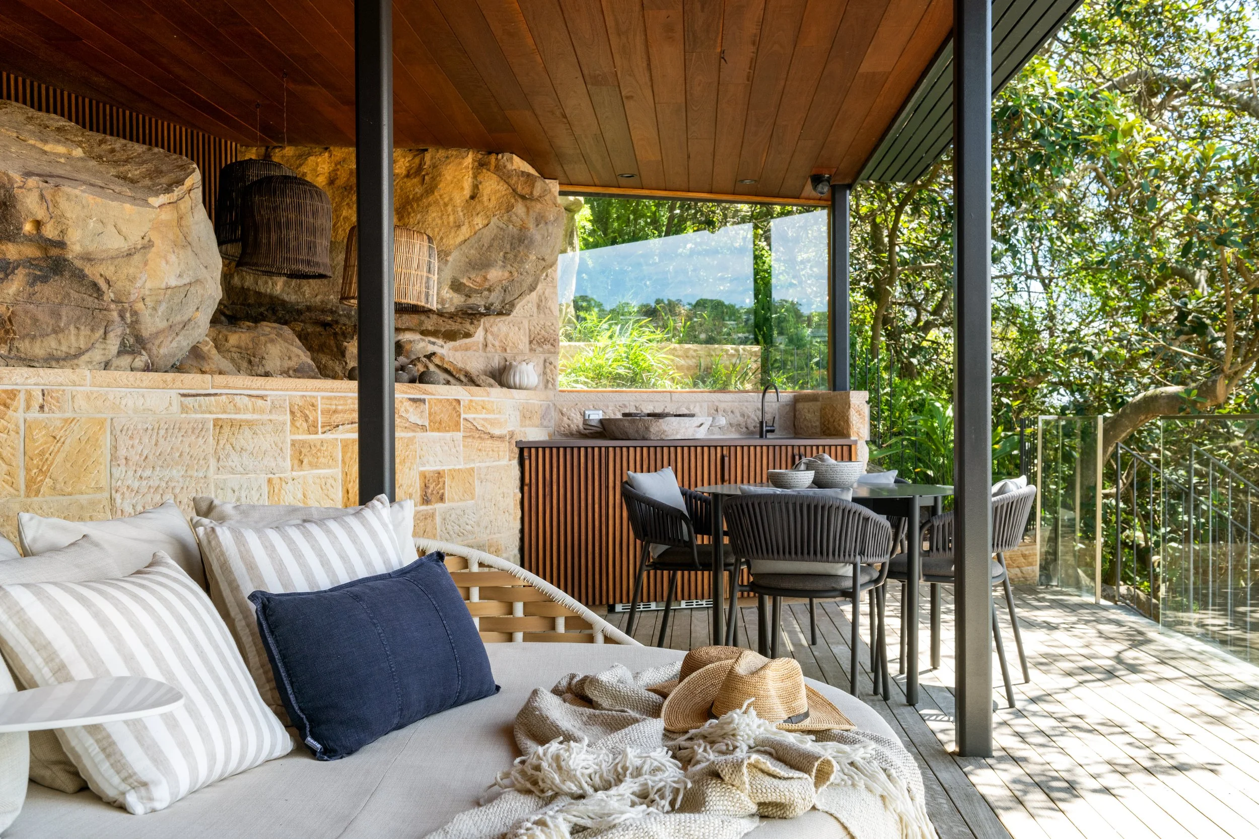 Outdoor living space with a beige striped sofa, pillows, a straw hat, and a blanket, opening to a patio with a dining table and chairs, surrounded by trees and greenery.