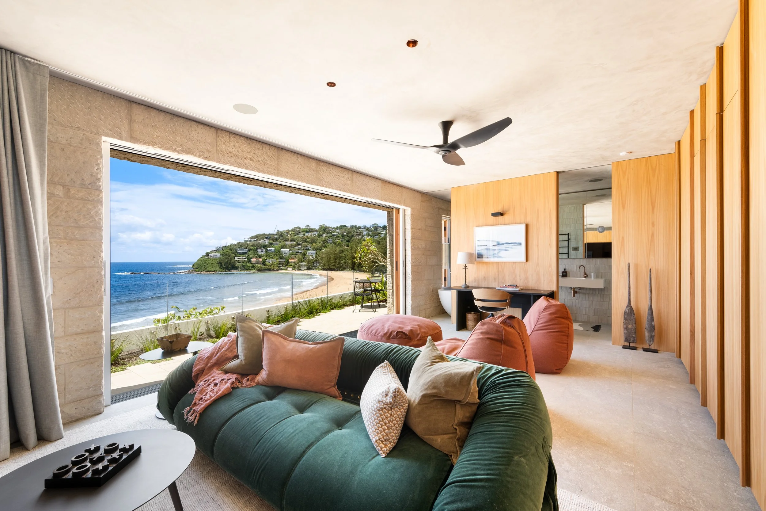 Living room with large open window showing ocean view, beige walls, green velvet sofa with pillows, wooden wall panels, ceiling fan, framed artwork, side table with lamp, and a sliding door leading to the outdoor beach area.
