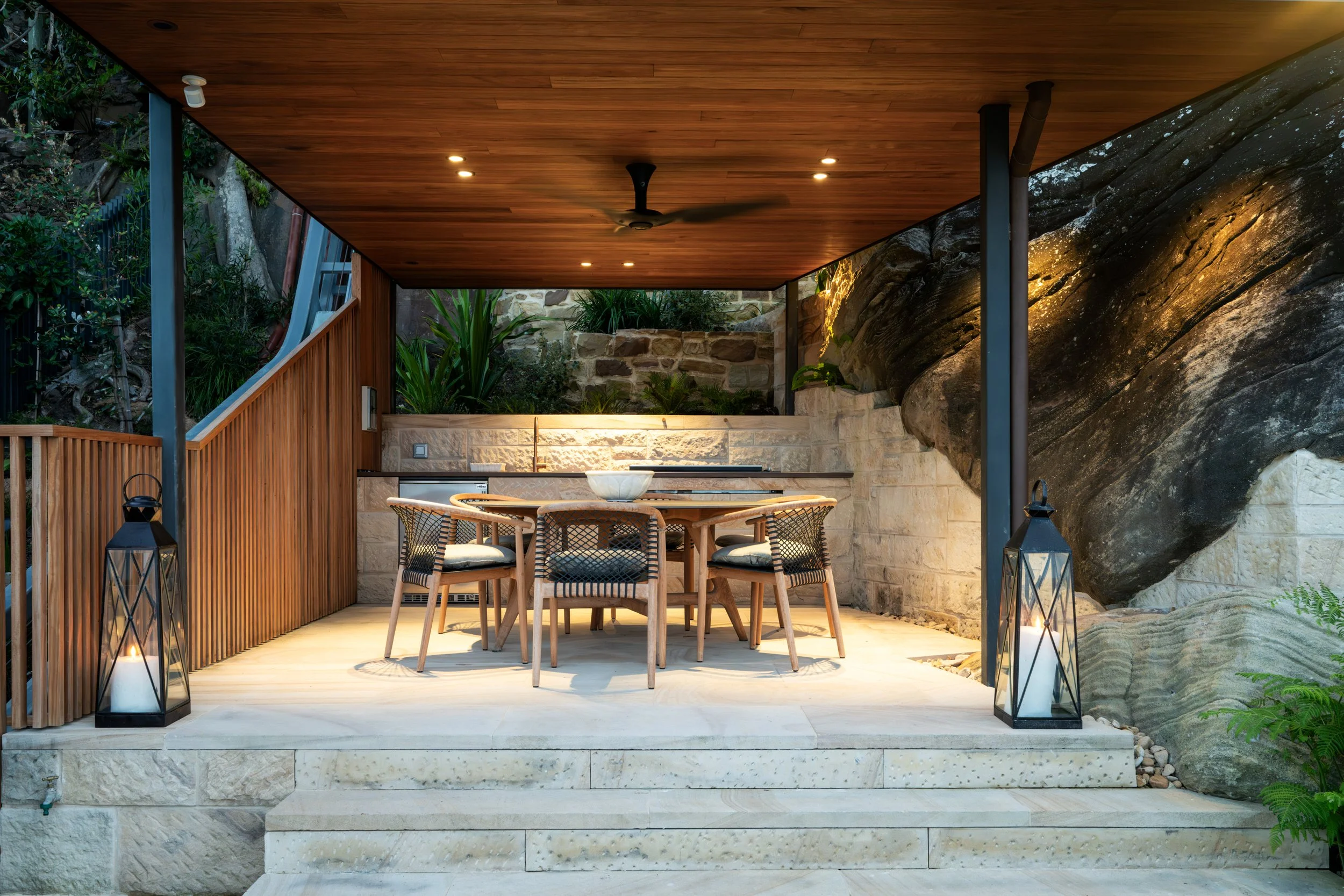 Outdoor dining area with wooden ceiling and stone walls, surrounded by rocks and greenery, featuring a round wooden table with chairs, lanterns, and ambient lighting.