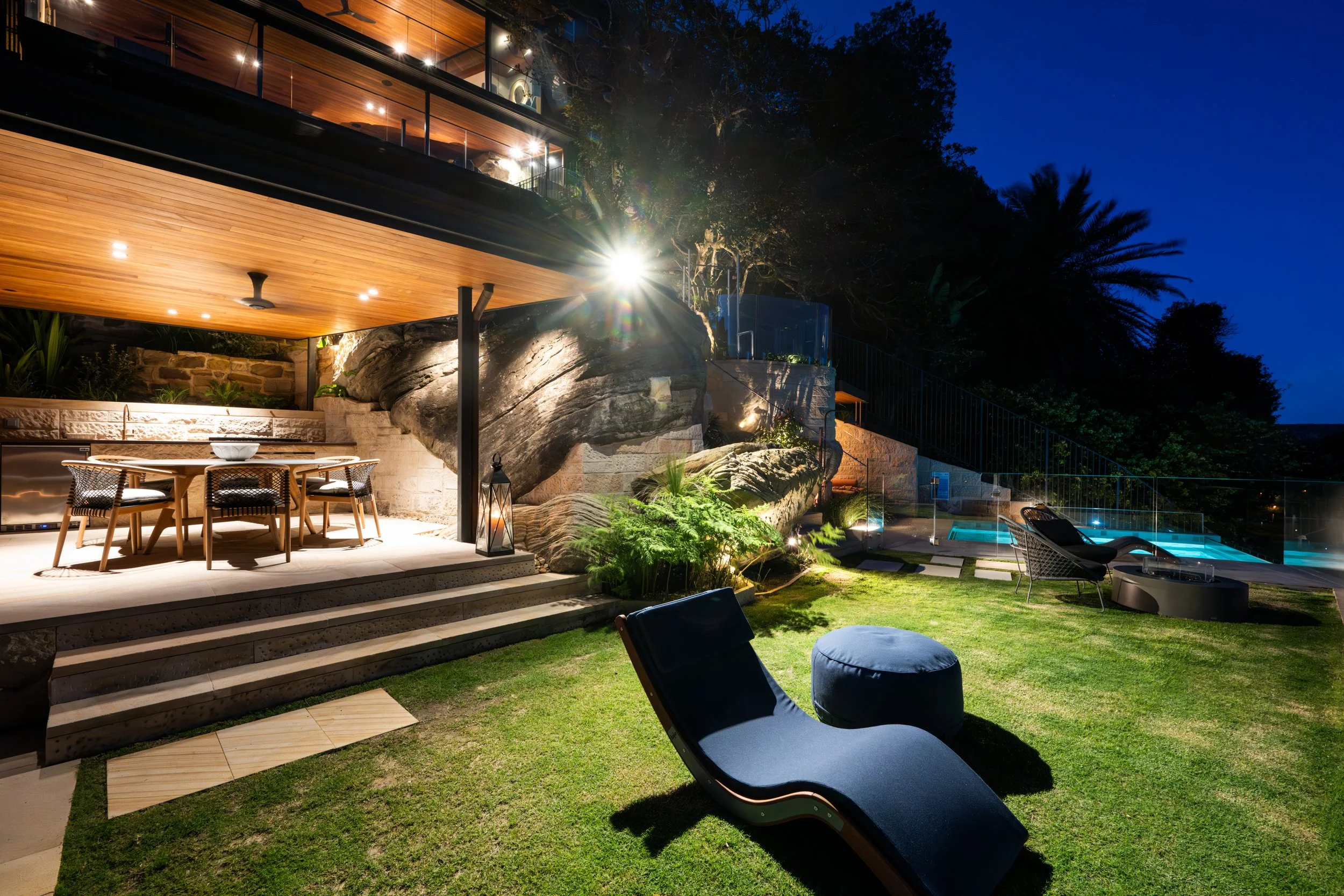 Night view of a modern backyard with a seating area, illuminated rocks and plants, a pool, and a multi-level house with wooden and glass exterior.