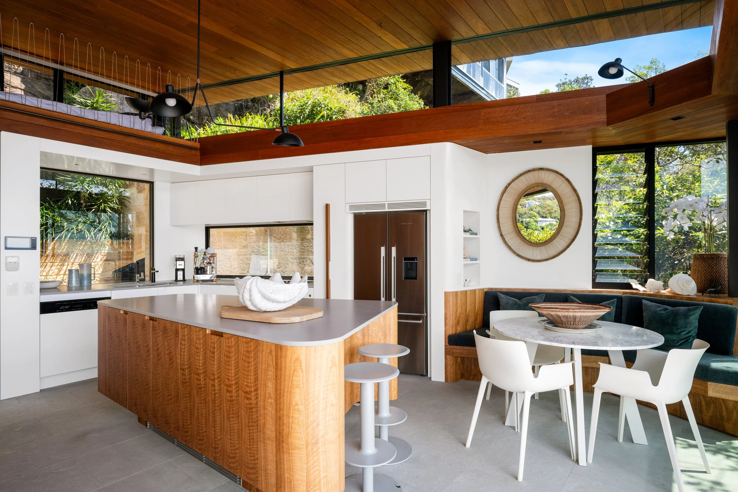 Open-concept kitchen and living area with wooden ceiling, white cabinets, marble-topped island, round dining table with white chairs, large windows with lush greenery outside, and wall decor.
