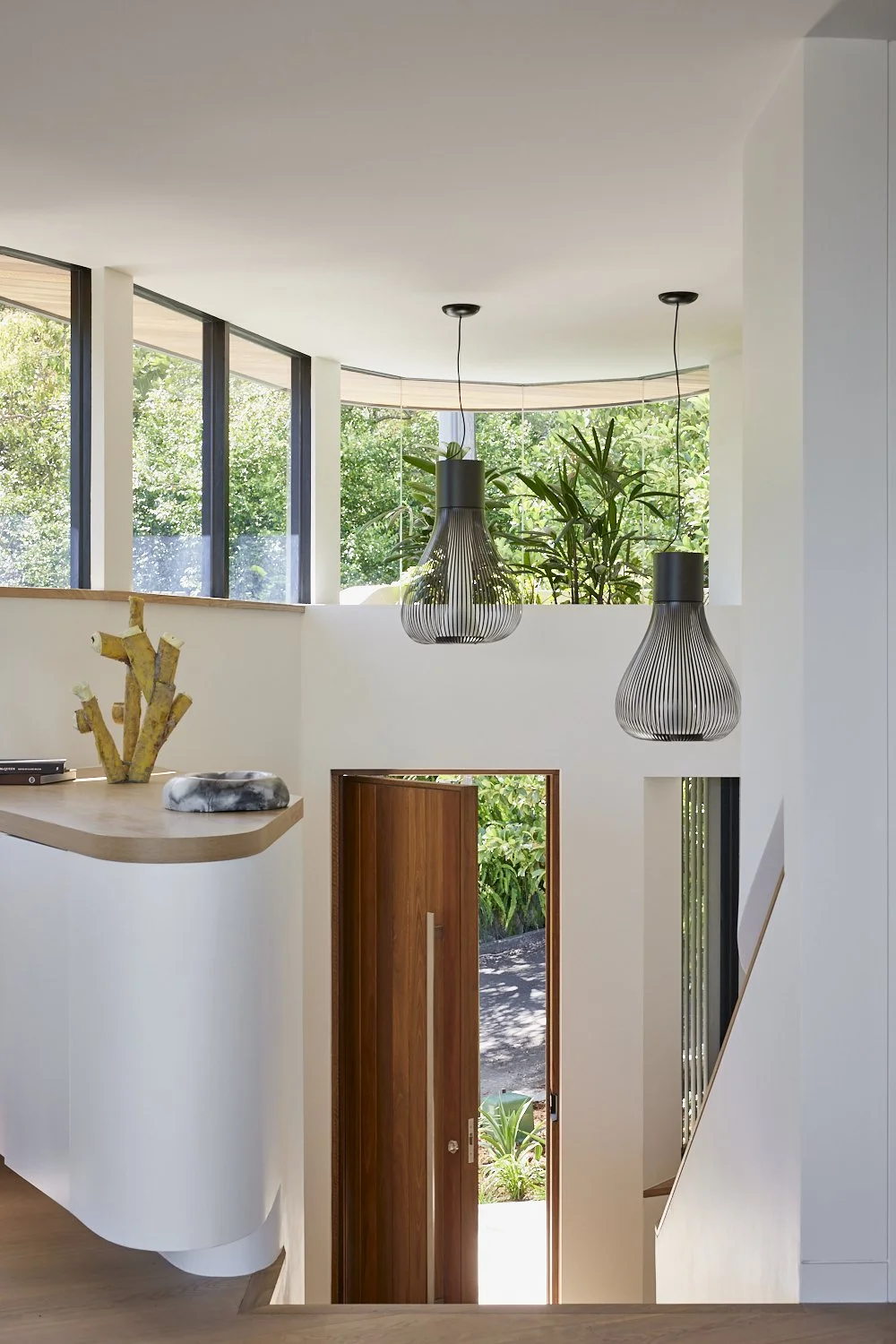 Interior view of a modern house with large windows, green plants, and hanging black wire pendant lights near a wooden door.