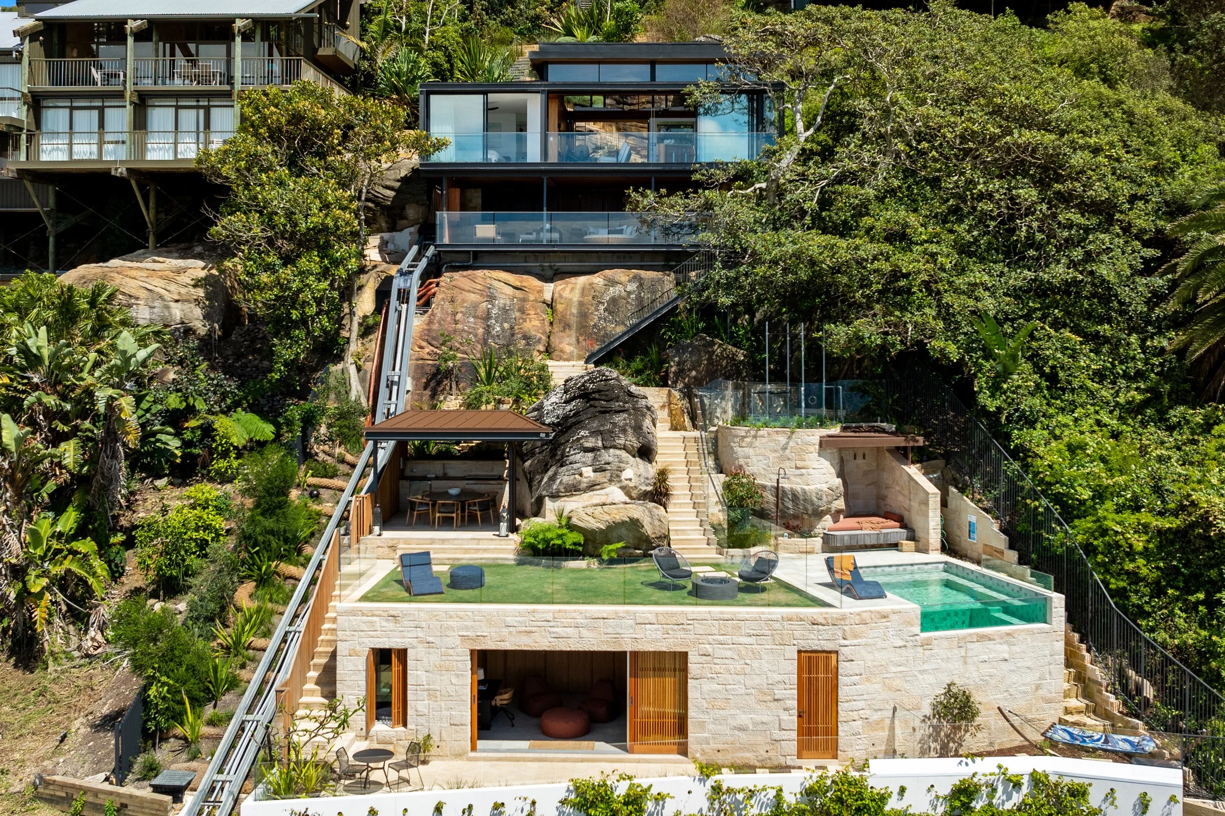 Multi-level modern house built into a hillside, featuring glass balconies, outdoor seating, pool area with lounge chairs, surrounded by lush greenery and rocks.