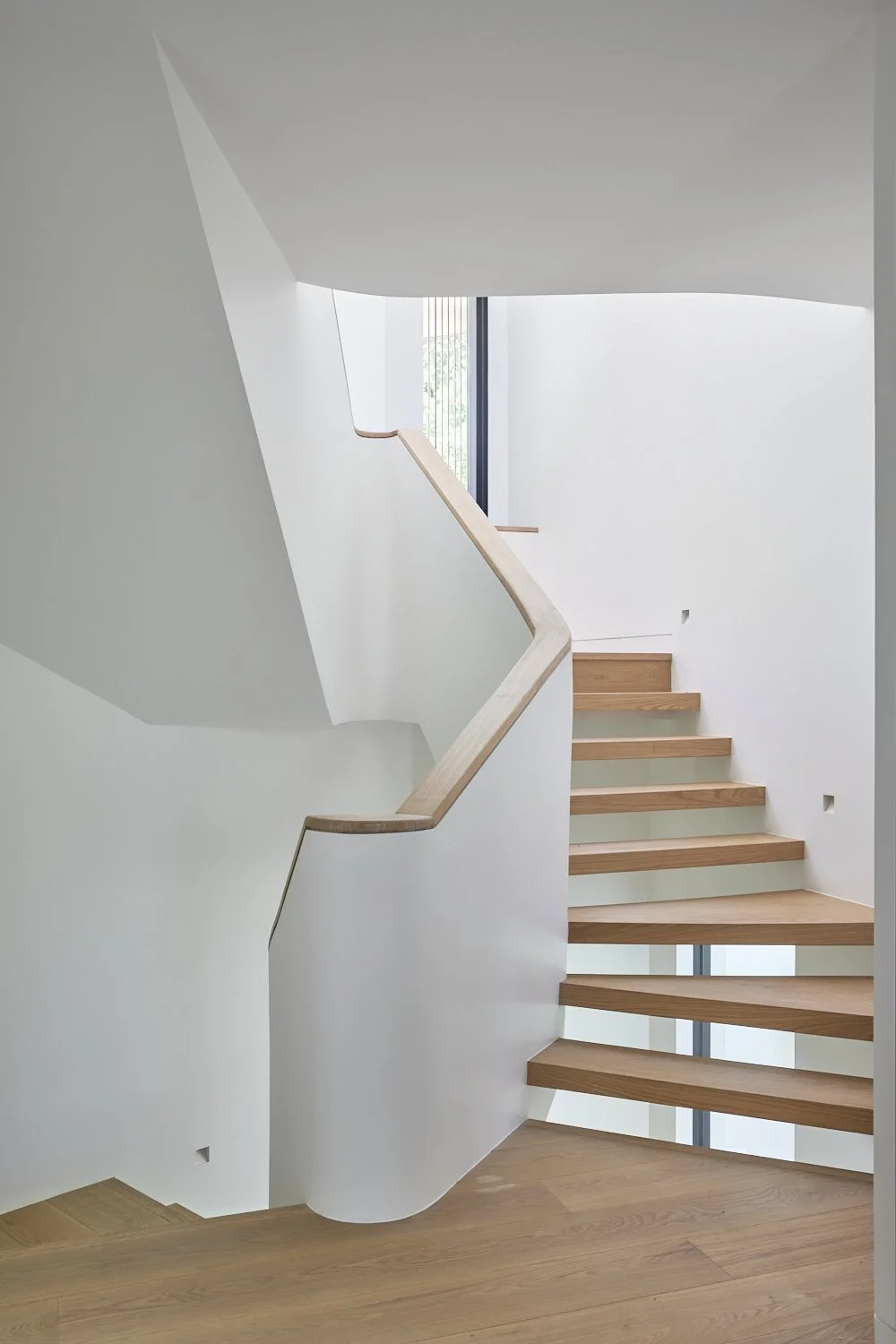 Modern interior staircase with wooden steps and a wooden handrail, white walls, and a large window at the top allowing natural light.