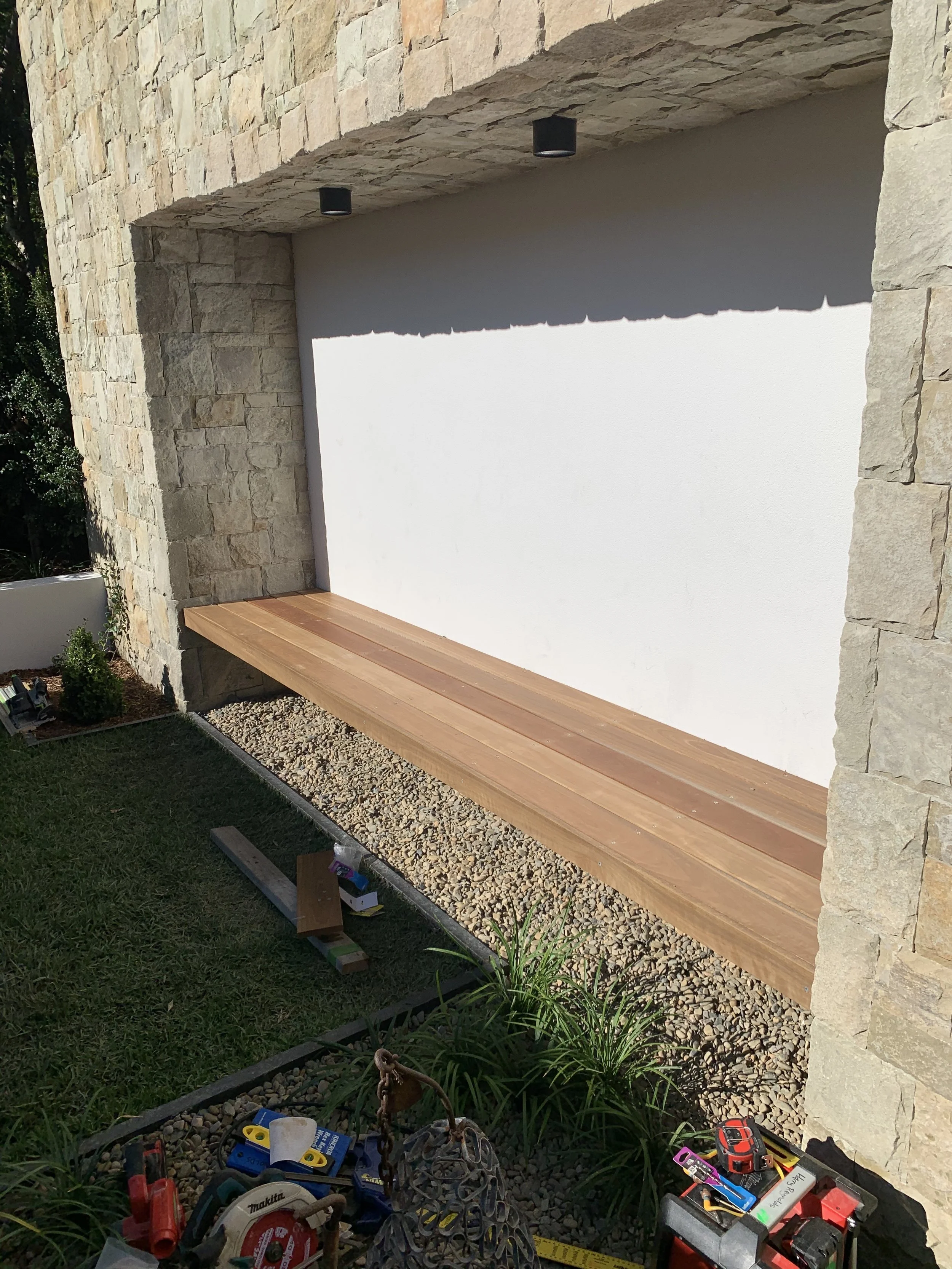 A construction site with a stone wall, a wooden bench, tools, and a grassy area with small plants.
