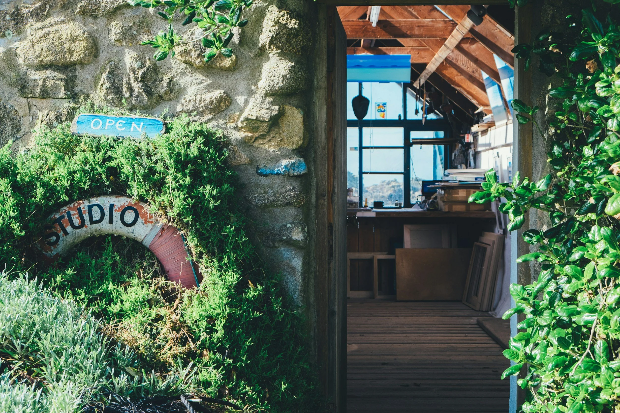 A rustic outdoor scene with a stone wall partially covered in green plants and a small blue sign that says 'OPEN'. To the right, there is an opening into a wooden structure with large windows allowing natural light, with some shelves, artwork, and various items visible inside.