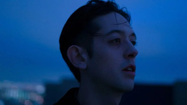 A young person with short dark hair looking into the distance against a dusky blue sky.