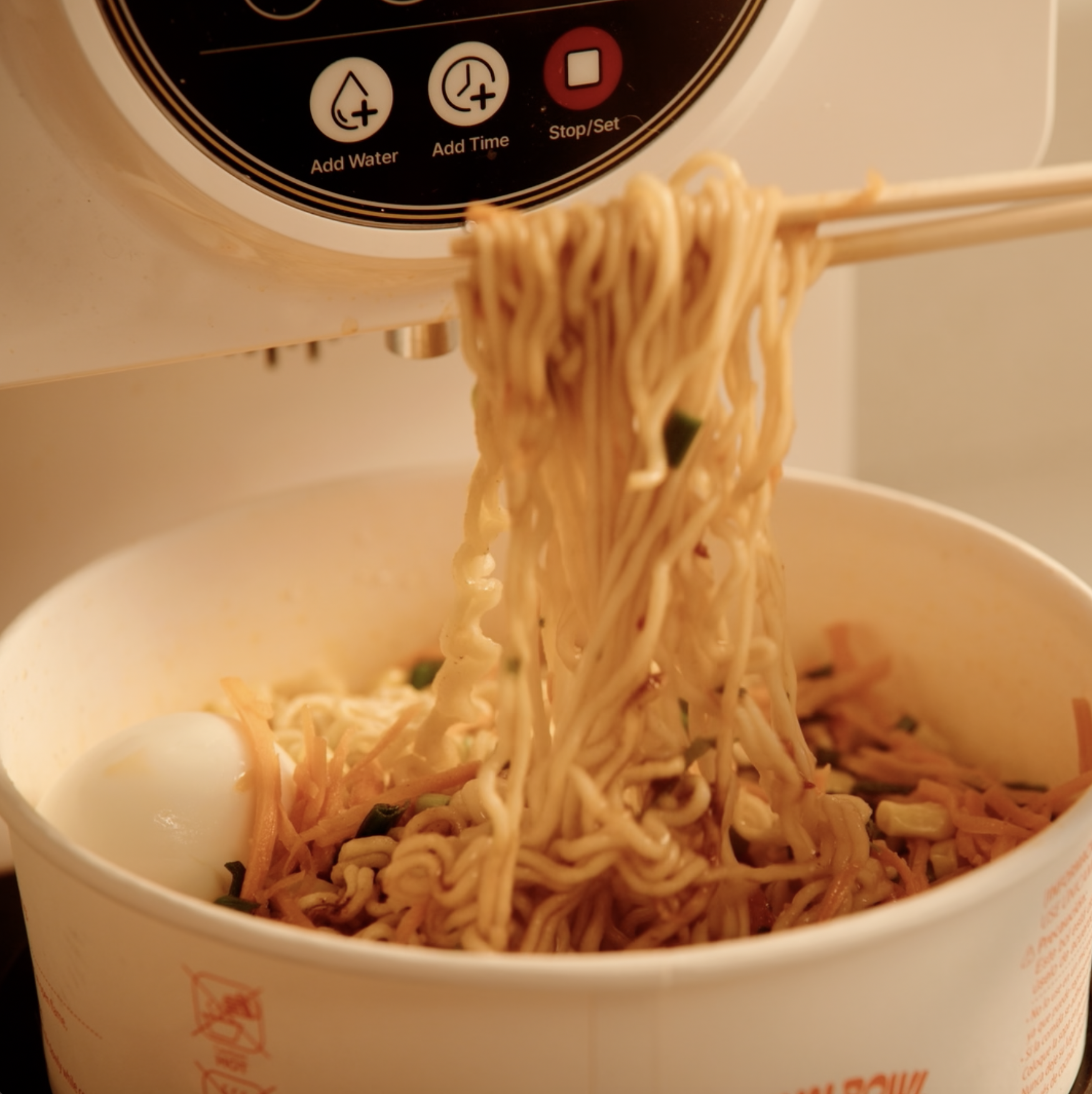 Close-up of a cup of ramen noodles with a soft-boiled egg and vegetables, with part of a rice cooker control panel visible in the background.