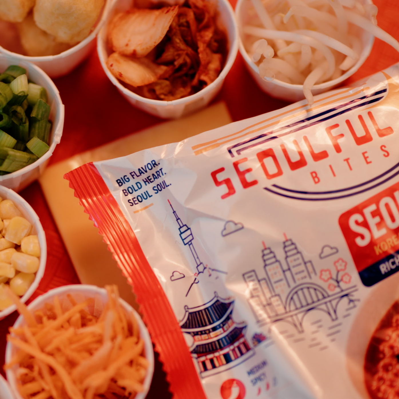 Close-up of a bag of Seoulful Bites with various small bowls of Asian-inspired side dishes or toppings around it, including chopped green onions, shredded carrots, bean sprouts, corn, and kimchi.