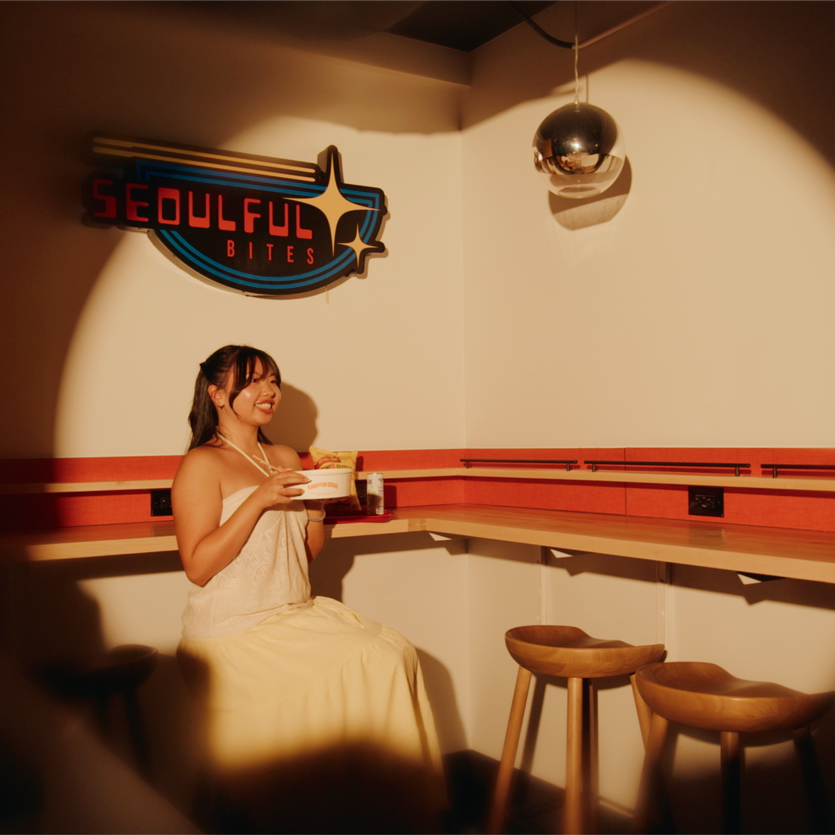 A woman wearing a beige dress sitting in a cozy room with a bowl in her hands, illuminated by warm lighting. There is a sign on the wall that reads 'Soulful Bites' and modern wooden stools nearby.