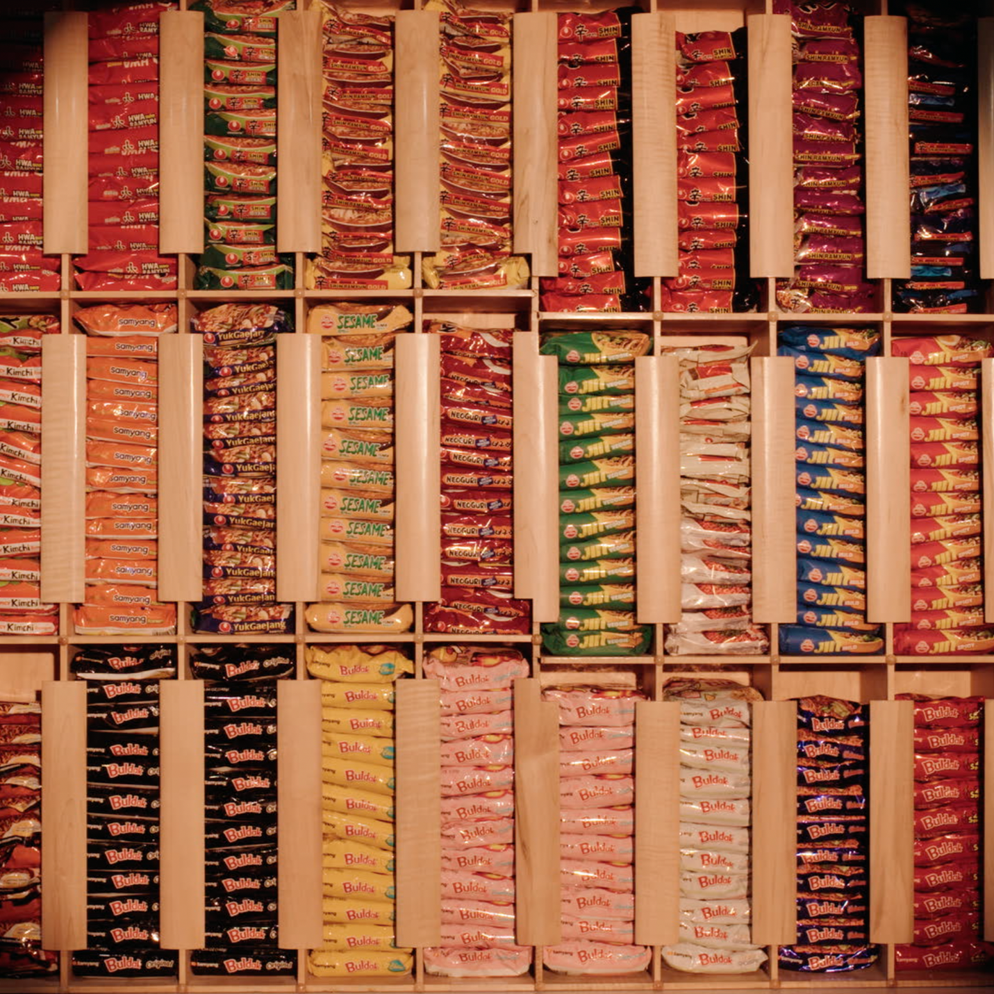 A wall of neatly organized instant noodle packages in various colors, stacked in compartments inside a wooden shelf.