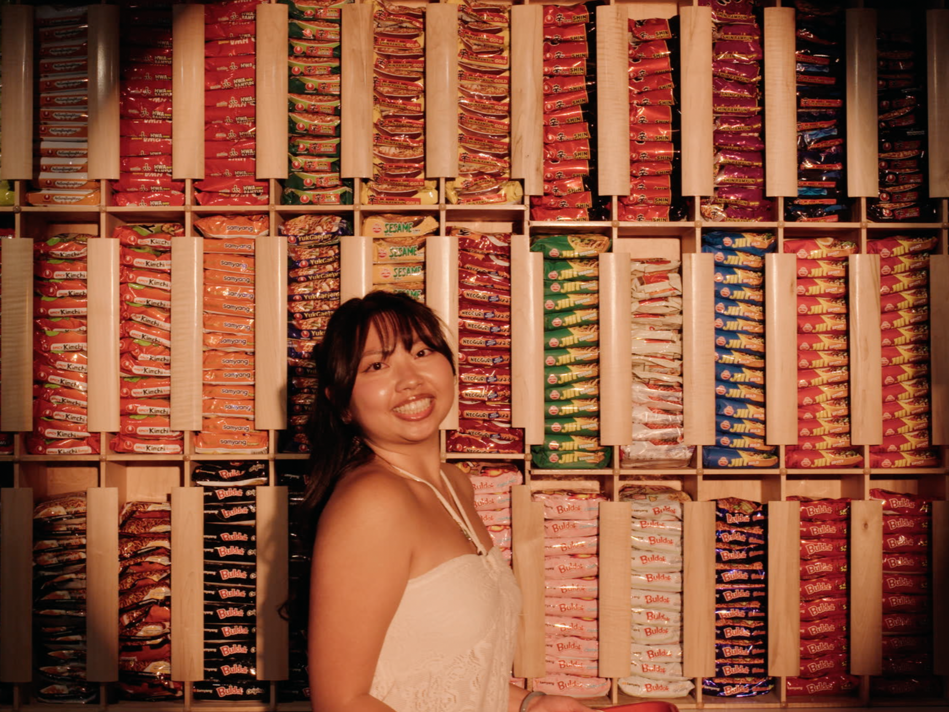 A woman smiling and standing in front of a large wall of many colorful snack packages stacked in compartments.