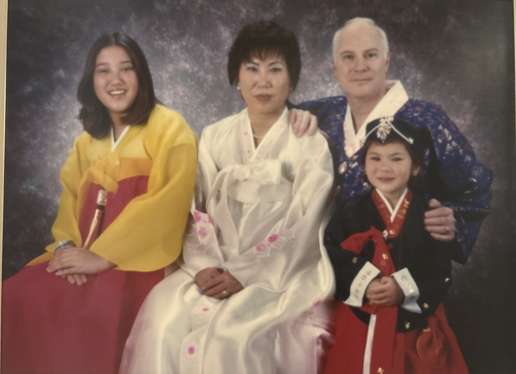 A group of five people dressed in traditional Korean hanbok, posing for a formal family portrait against a dark, textured background. The group includes a young girl in a black dress with red accents, a woman in a pale pink hanbok with floral embroidery, an older woman in yellow, and an older man in a blue and gold patterned hanbok, with a younger woman in pink and red seated in front.
