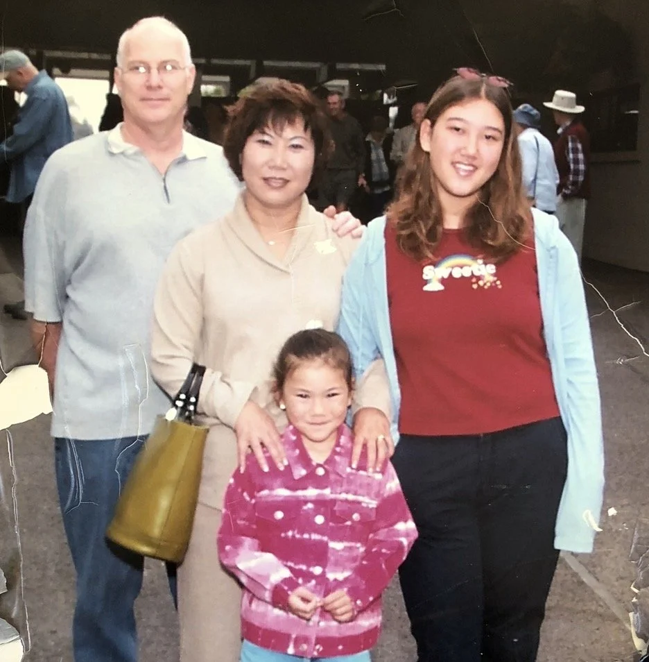 Group photo of four people, two adults and two children, standing indoors at a gathering or event. The man on the left has white hair and glasses, wearing a light gray shirt. The woman in the middle has short dark hair, wearing a beige top. The girl on the right has long brown hair, wearing a red shirt with a rainbow graphic and a light blue jacket. The young girl in front has dark hair, wearing a pink and white tie-dye jacket, smiling at the camera.