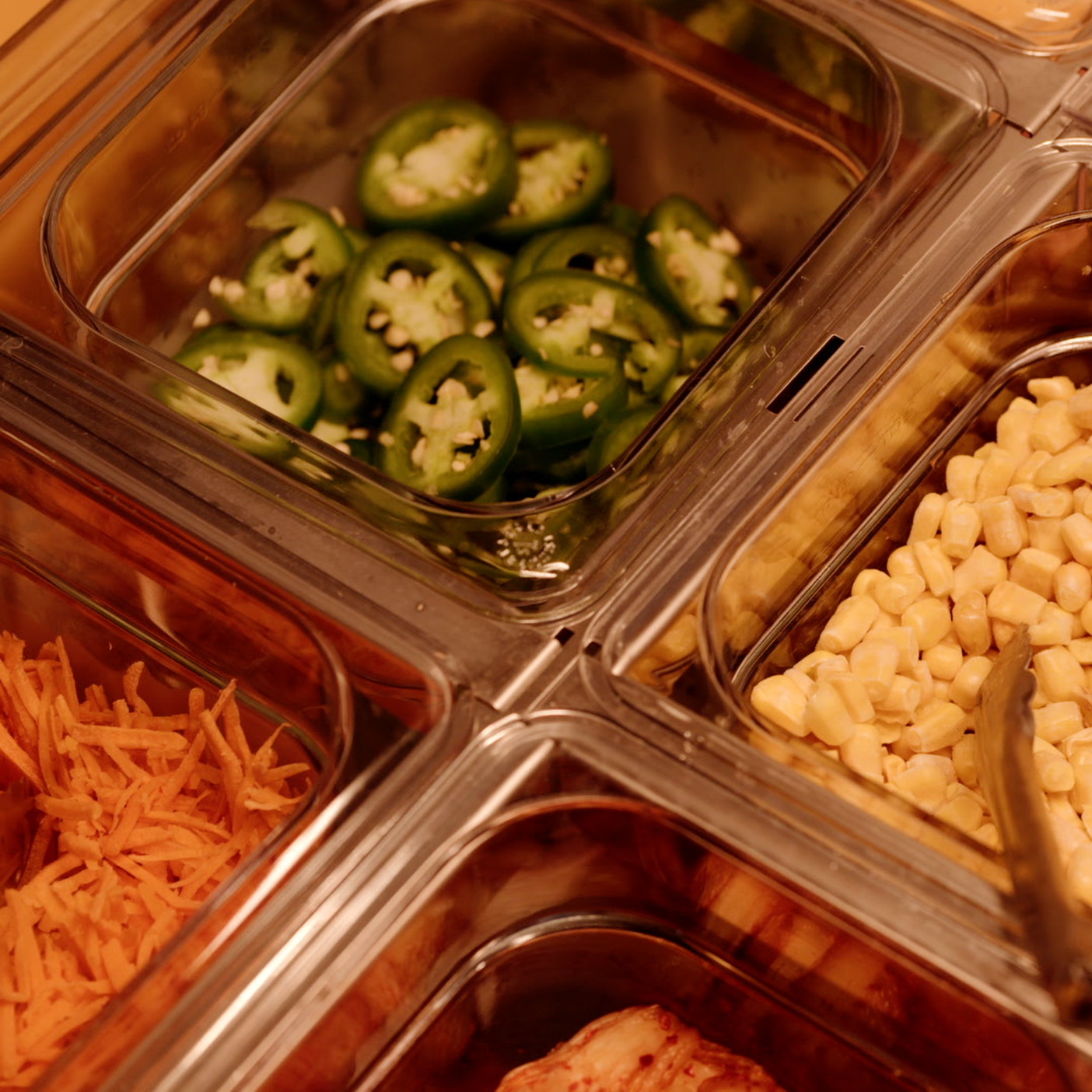 Close-up of a clear food storage container with sliced jalapeño peppers inside, surrounded by other food items in separate compartments such as shredded cheese, corn, and shredded carrots.
