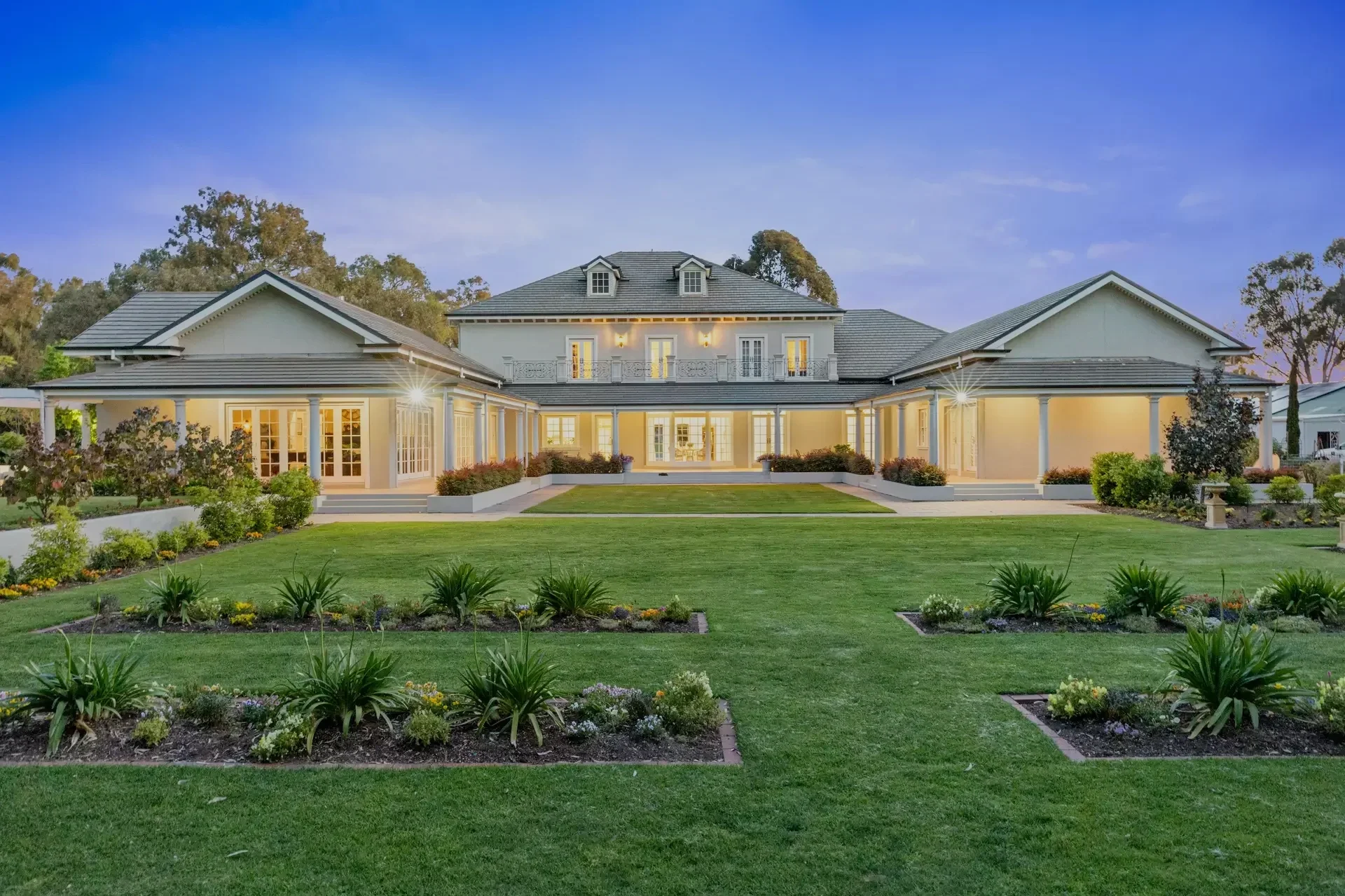 Large, elegant house with multiple gabled roofs, illuminated from inside, surrounded by a well-manicured lawn and landscaped garden with bushes and flowers, during dusk.