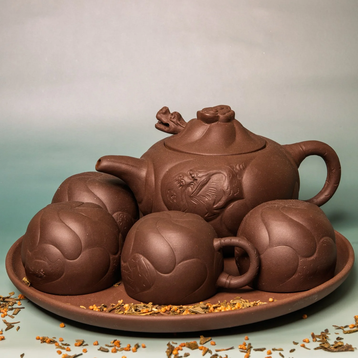 A Yixing purple-clay teapot with matching cups on a tray, scattered loose tea leaves alongside.