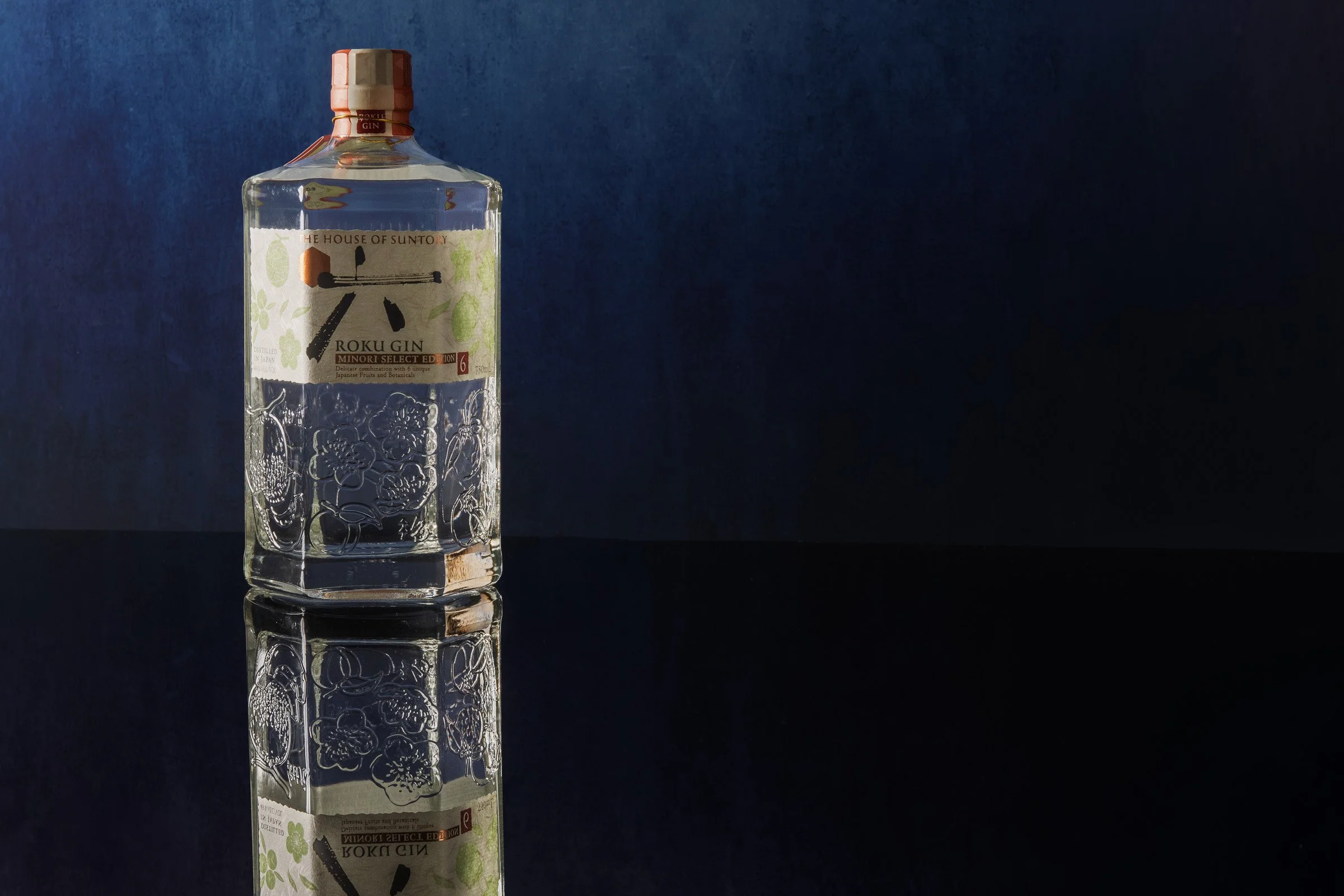 A bottle of Roku Japanese craft gin lit against a deep blue backdrop, with its reflection on a black surface.