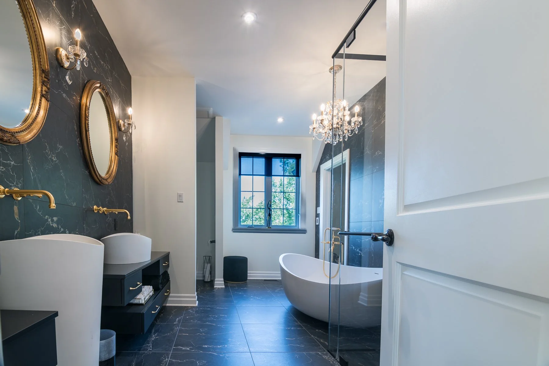 Primary bathroom in dark veined marble with gold oval mirrors, wall-mounted brass taps, and a freestanding tub