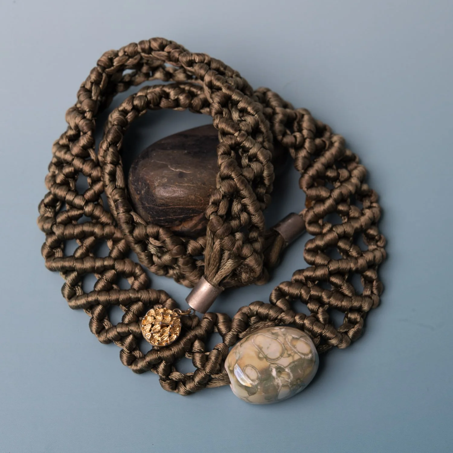 A knotted macrame necklace with river stone and carved beads on a pale blue surface.