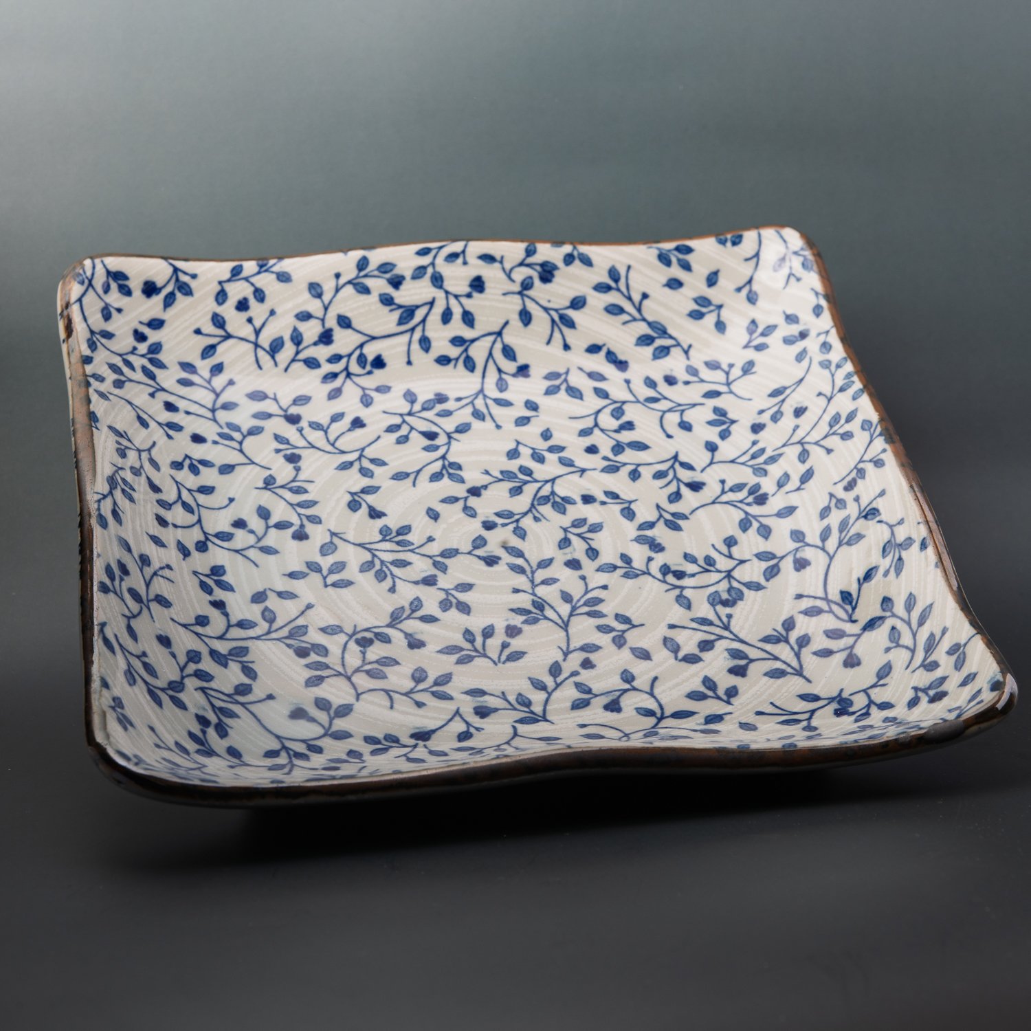 A square ceramic plate with wavy edges, hand-painted with a dense blue floral vine pattern on cream, against a dark grey backdrop.