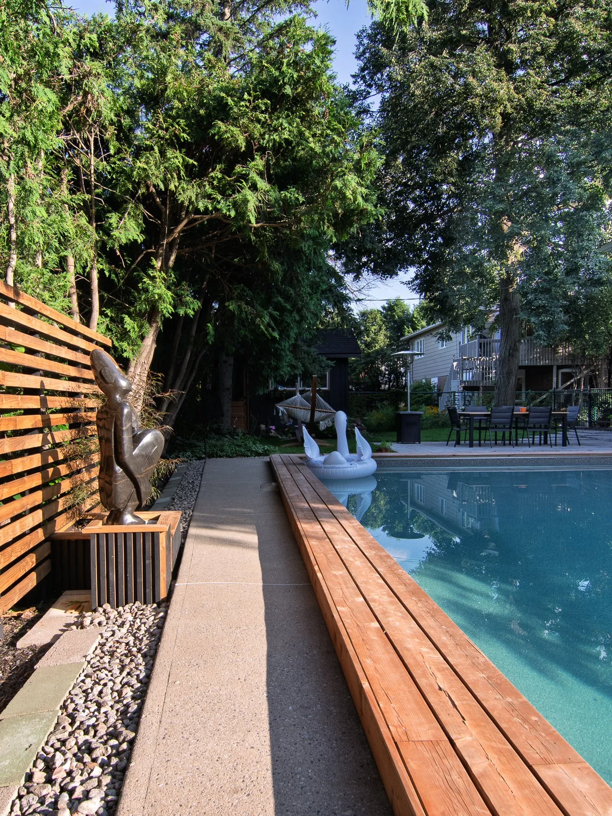 Grounds Cedar slat screen with a bronze figure on a plinth, beside a gravel bed and a concrete walkway in late afternoon light.