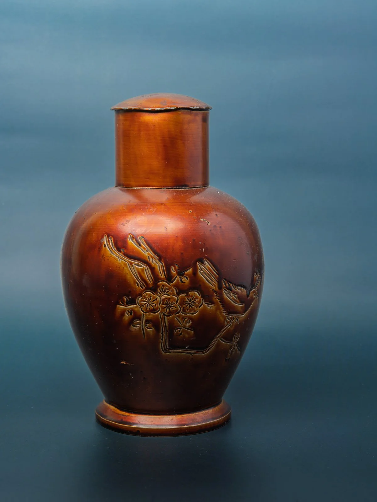 A copper-glazed ceramic vase with an incised design, lit against a blue-grey studio backdrop.