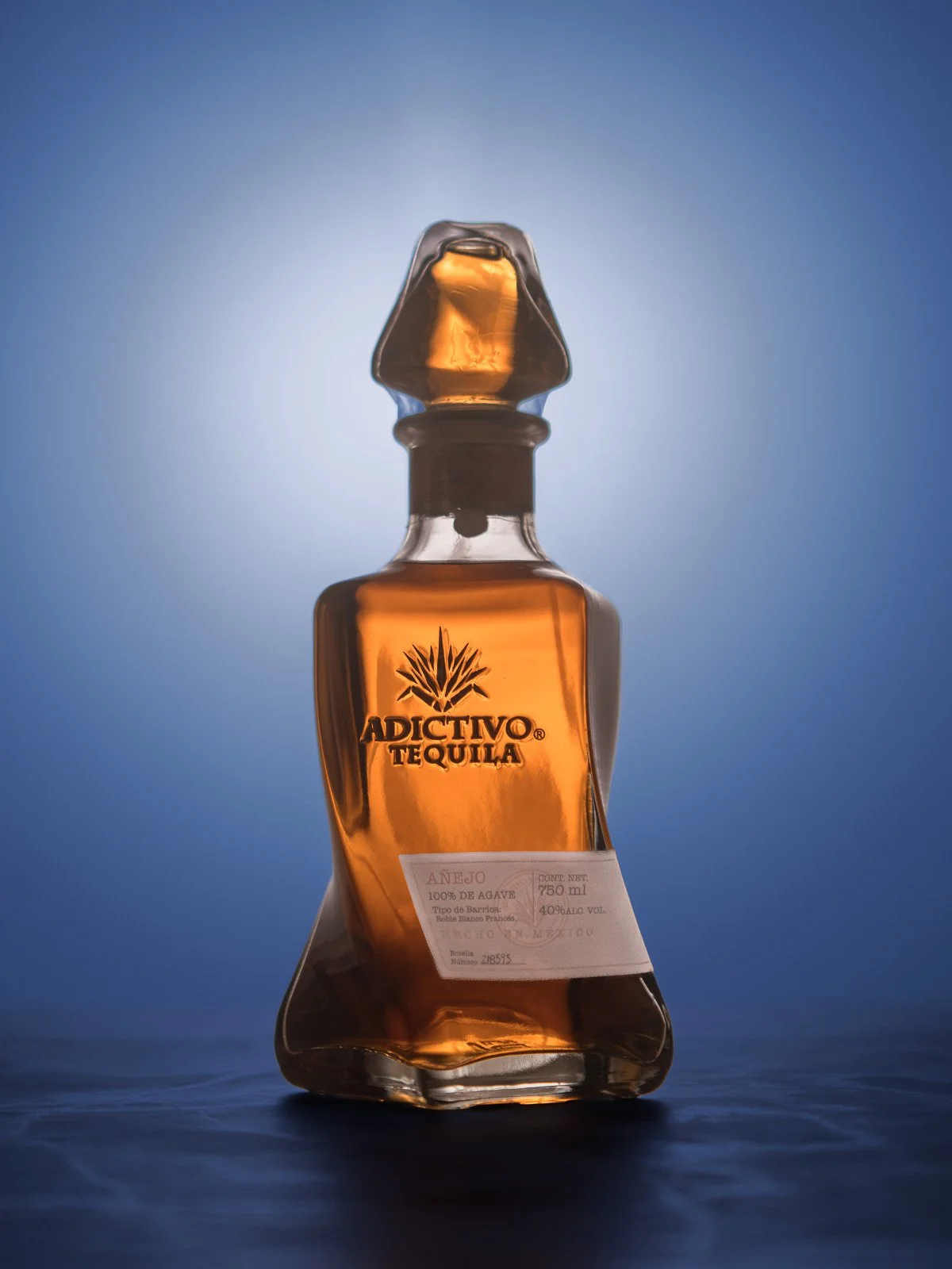 A bottle of Adictivo añejo tequila lit against a deep blue backdrop.