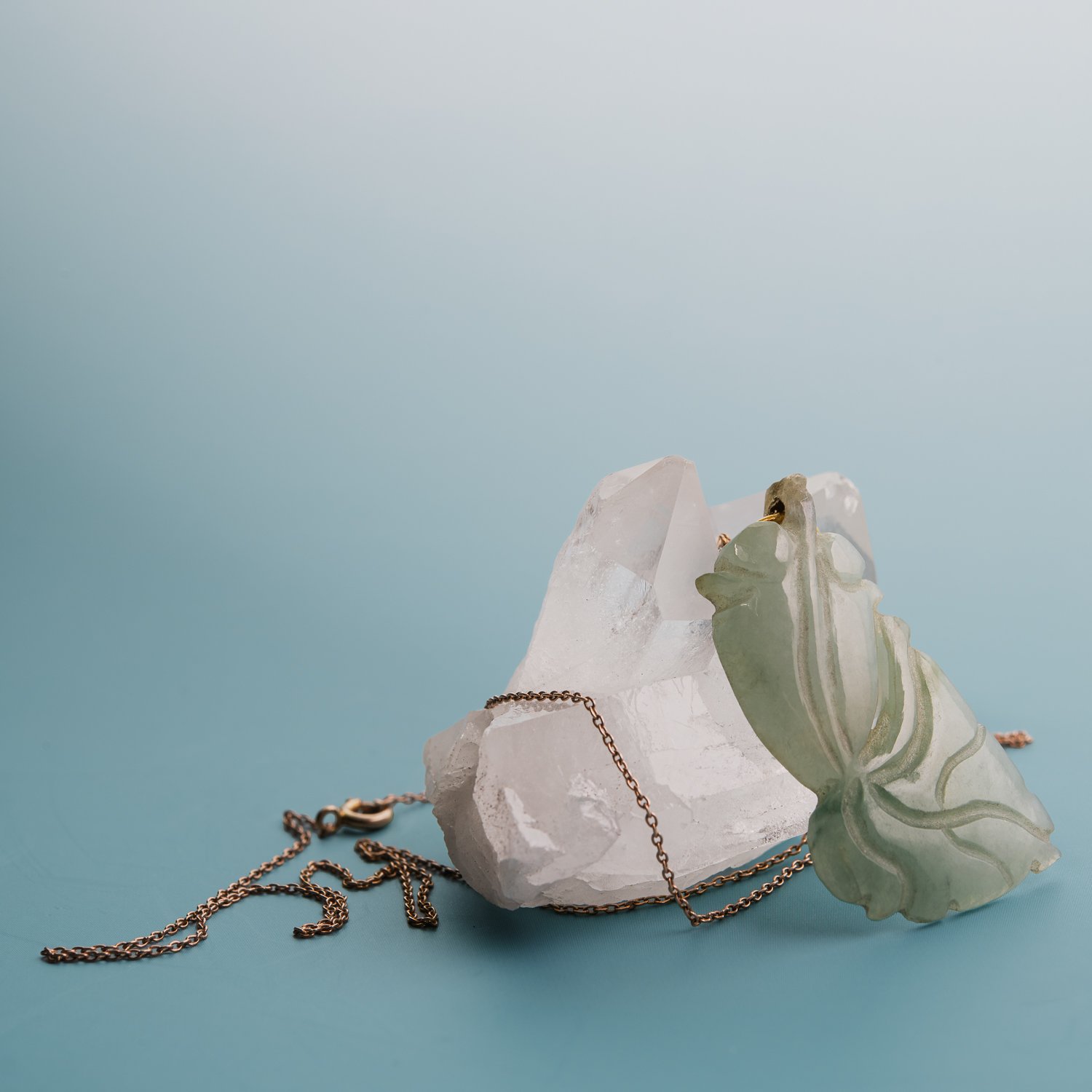 A carved pale green jade fish pendant hanging against a white quartz crystal, its gold chain pooling on a soft blue surface.