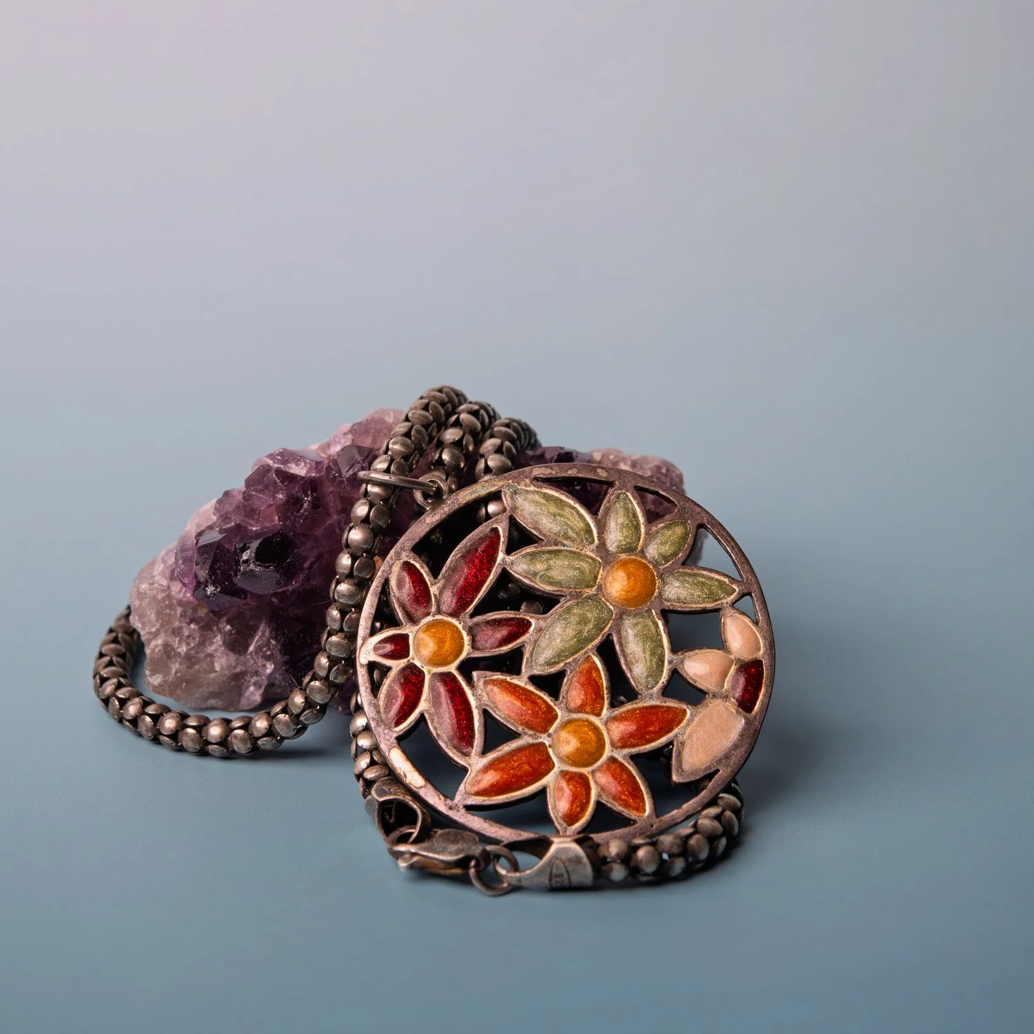 A round openwork silver pendant with enamelled flowers in red, green and orange, on a beaded chain, resting against an amethyst cluster.