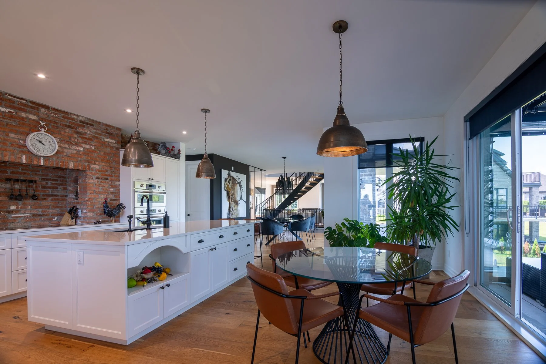 White kitchen with an exposed brick chimney breast, aged brass pendant lights, and a glass breakfast table