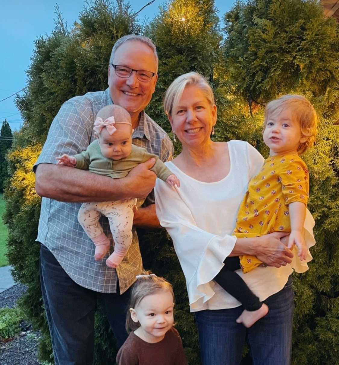 A smiling family of five posing outdoors at dusk with greenery in the background. An older man is holding a baby girl, a woman is holding another toddler girl, and a young girl stands in front, all appearing happy.
