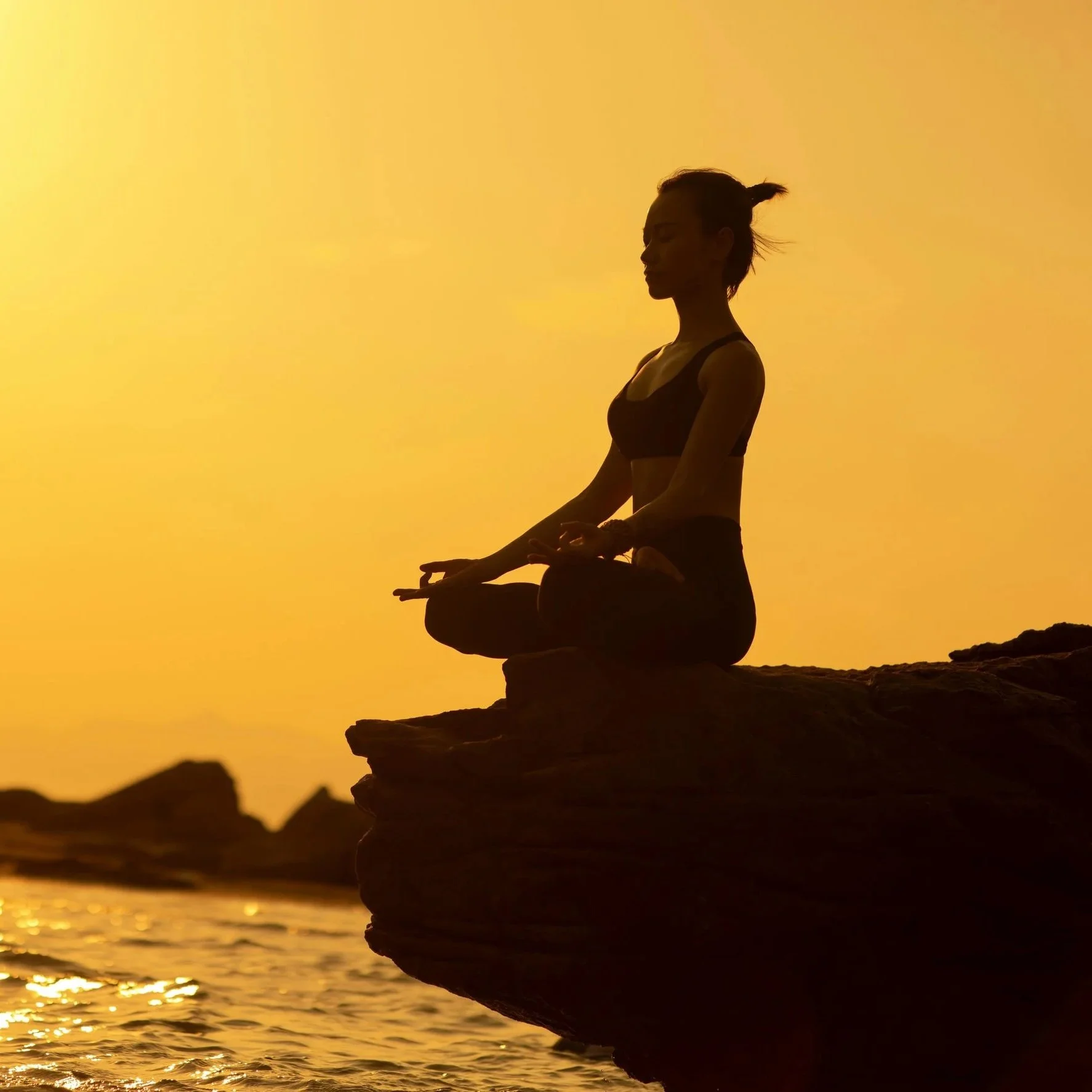 A person practicing yoga or meditation on a rock formation during sunset, with the ocean in the background.