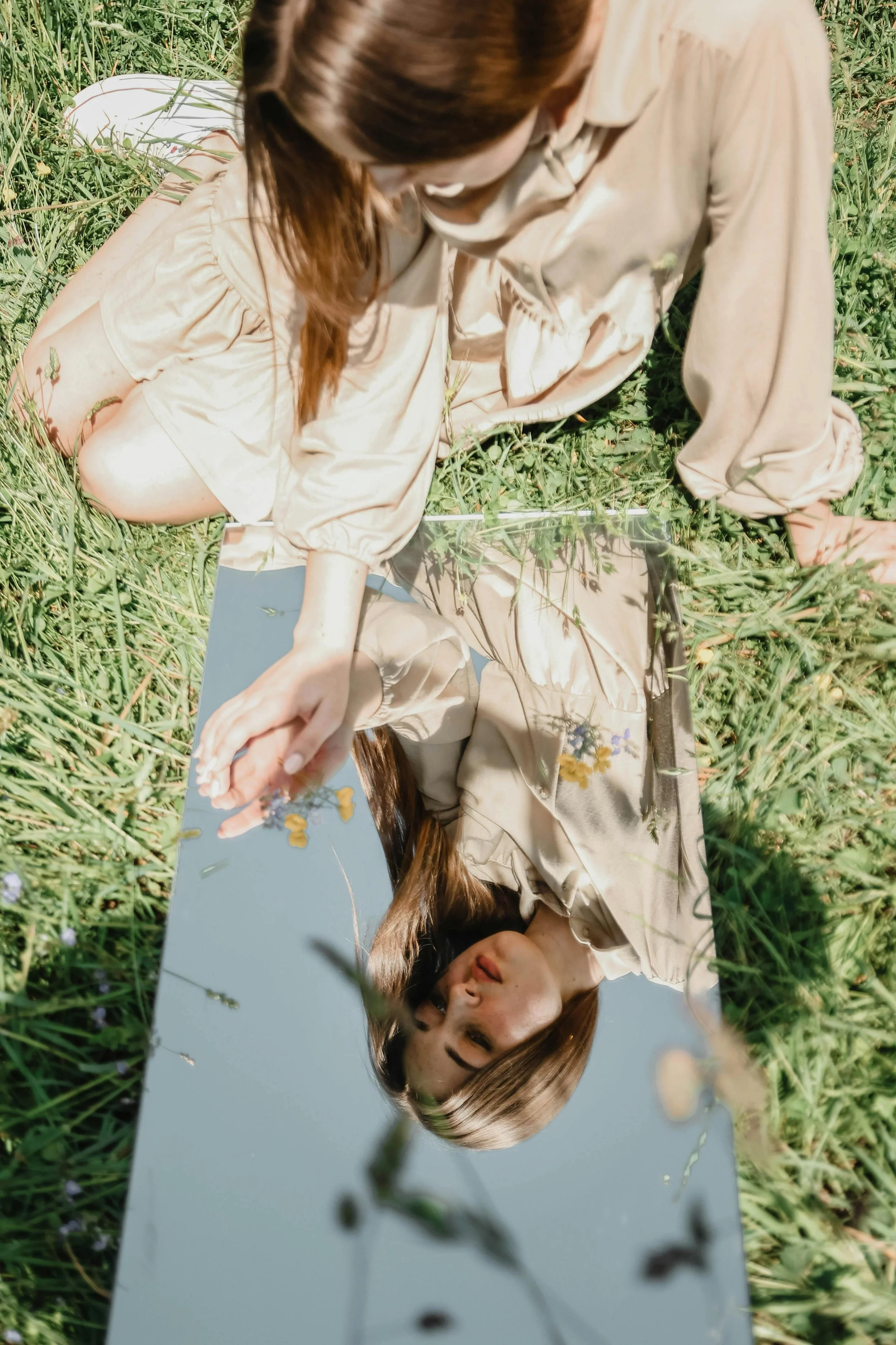 A woman with long hair wearing a beige dress is lying on a grassy field, looking at her reflection in a mirror placed on the ground.