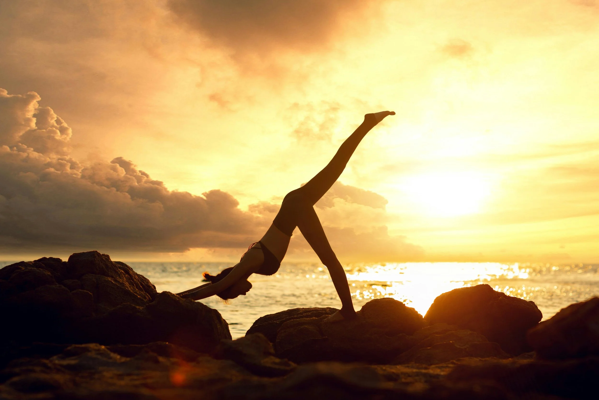 A person practicing yoga on rocks at sunset by the ocean.