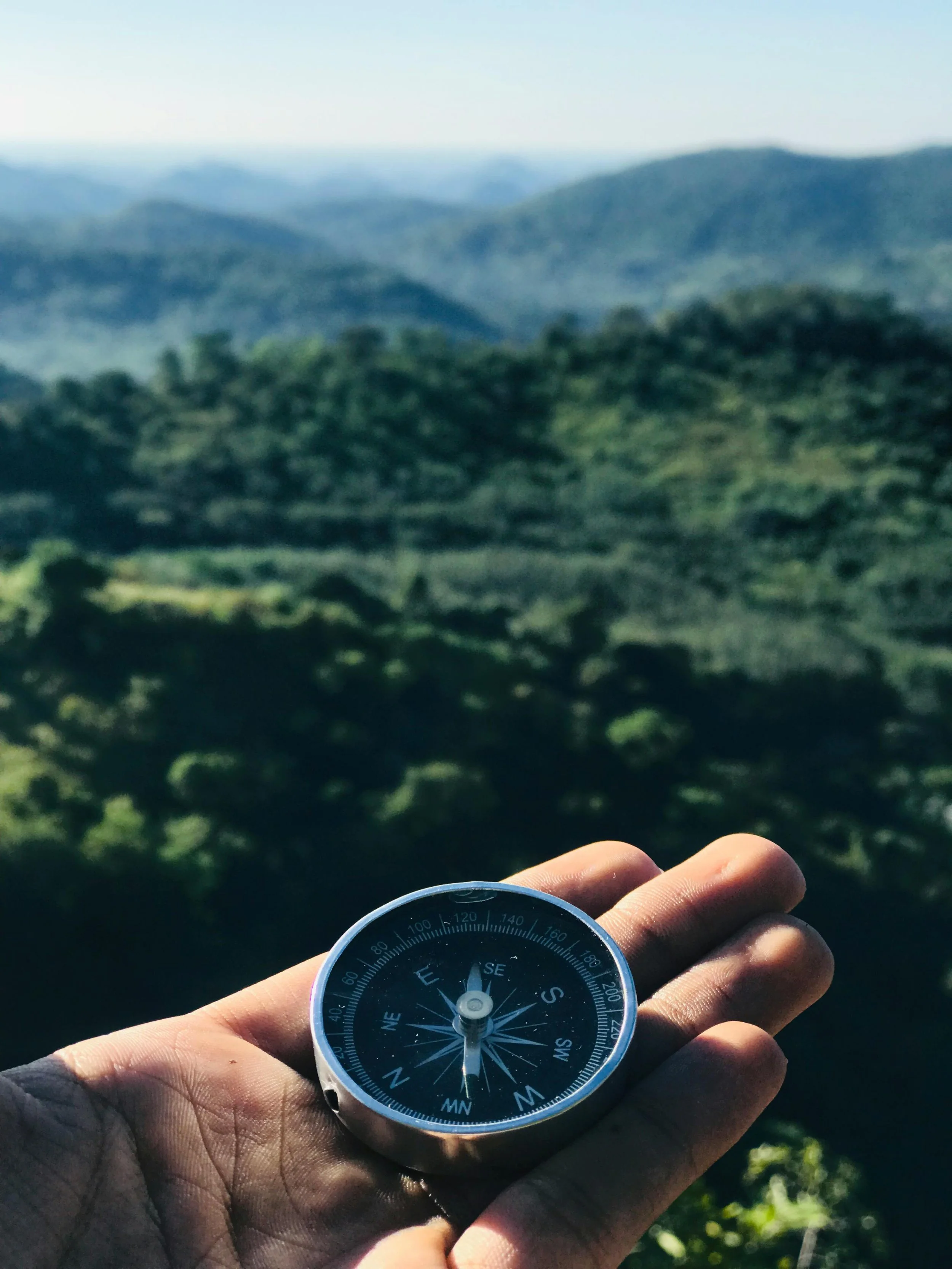 A person holding a compass outdoors overlooking a lush green landscape with rolling hills and distant mountains.