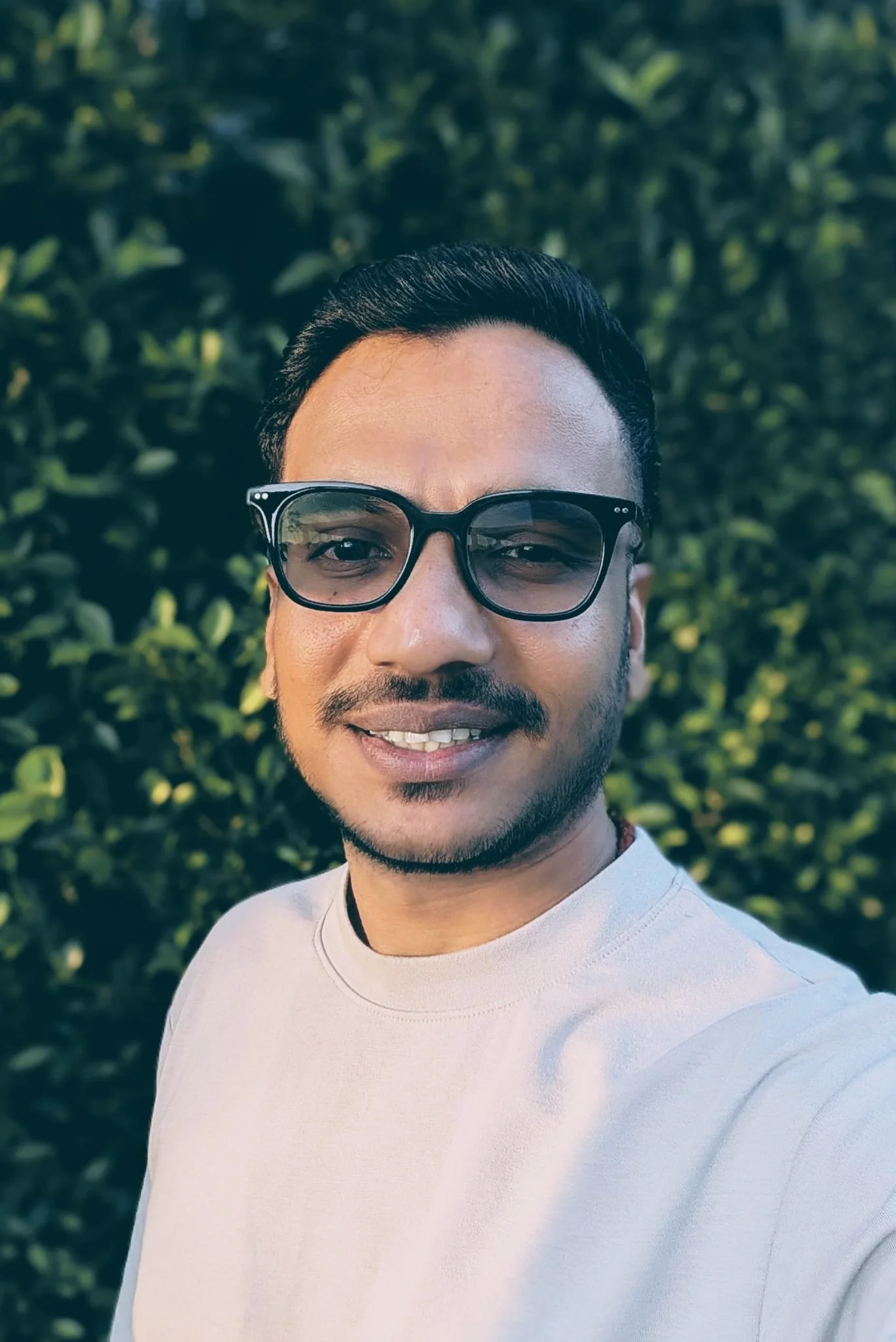 Close-up of a smiling man with glasses and short black hair, wearing a light gray shirt, standing outdoors in front of green bushes.