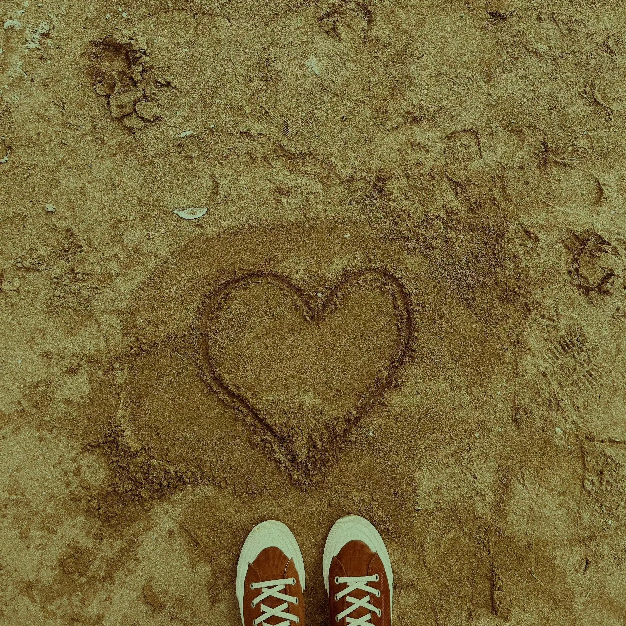 First person view of a person wearing red sneakers with white laces and white soles, standing on a sandy ground with footprints and tire tracks.