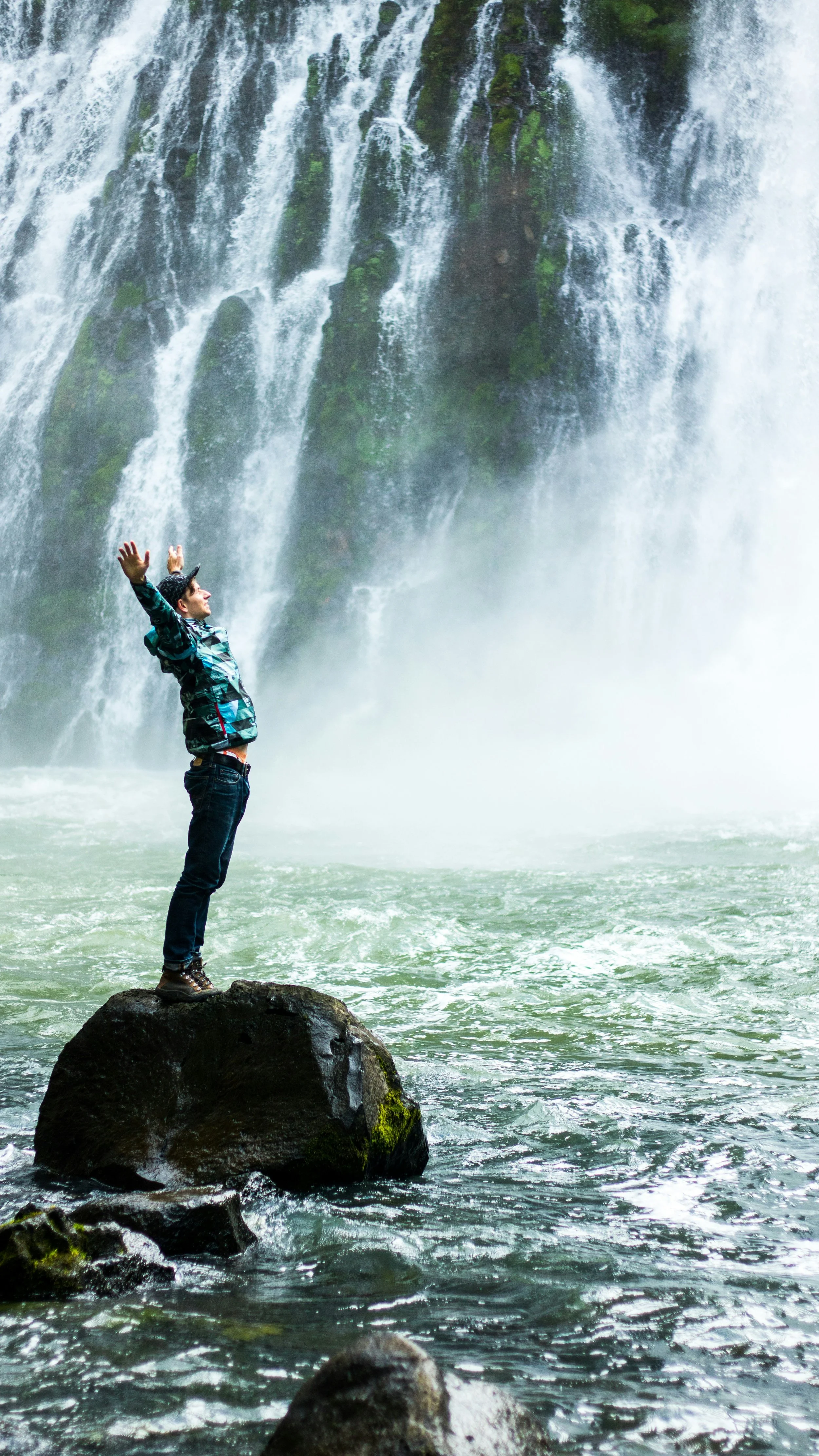 A person stands on a large rock in a river, with a waterfall in the background, arms raised, enjoying nature.