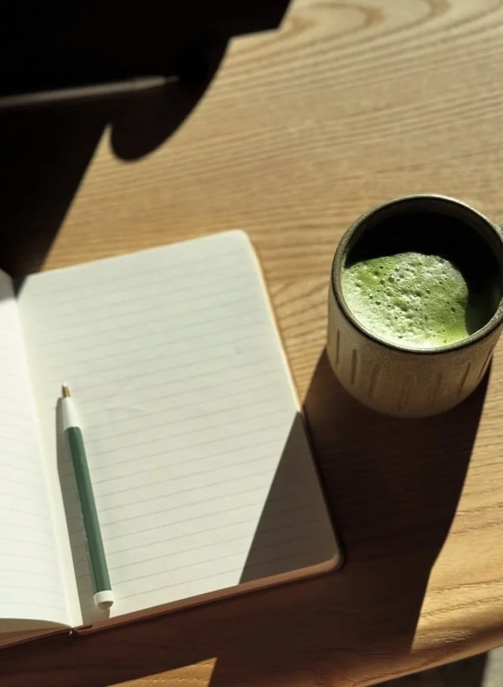 Cozy flat lay of a journal beside a cup of green matcha tea, set in soft natural lighting with a calm, minimal aesthetic