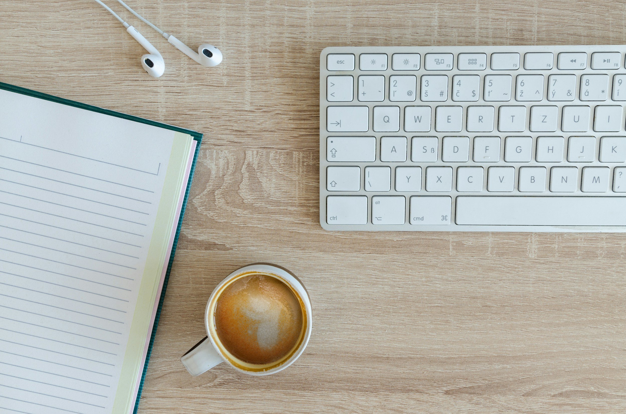 A wooden desk with white earphones, a notebook, a white keyboard, a cup of coffee, and a small jug of milk or cream on it.