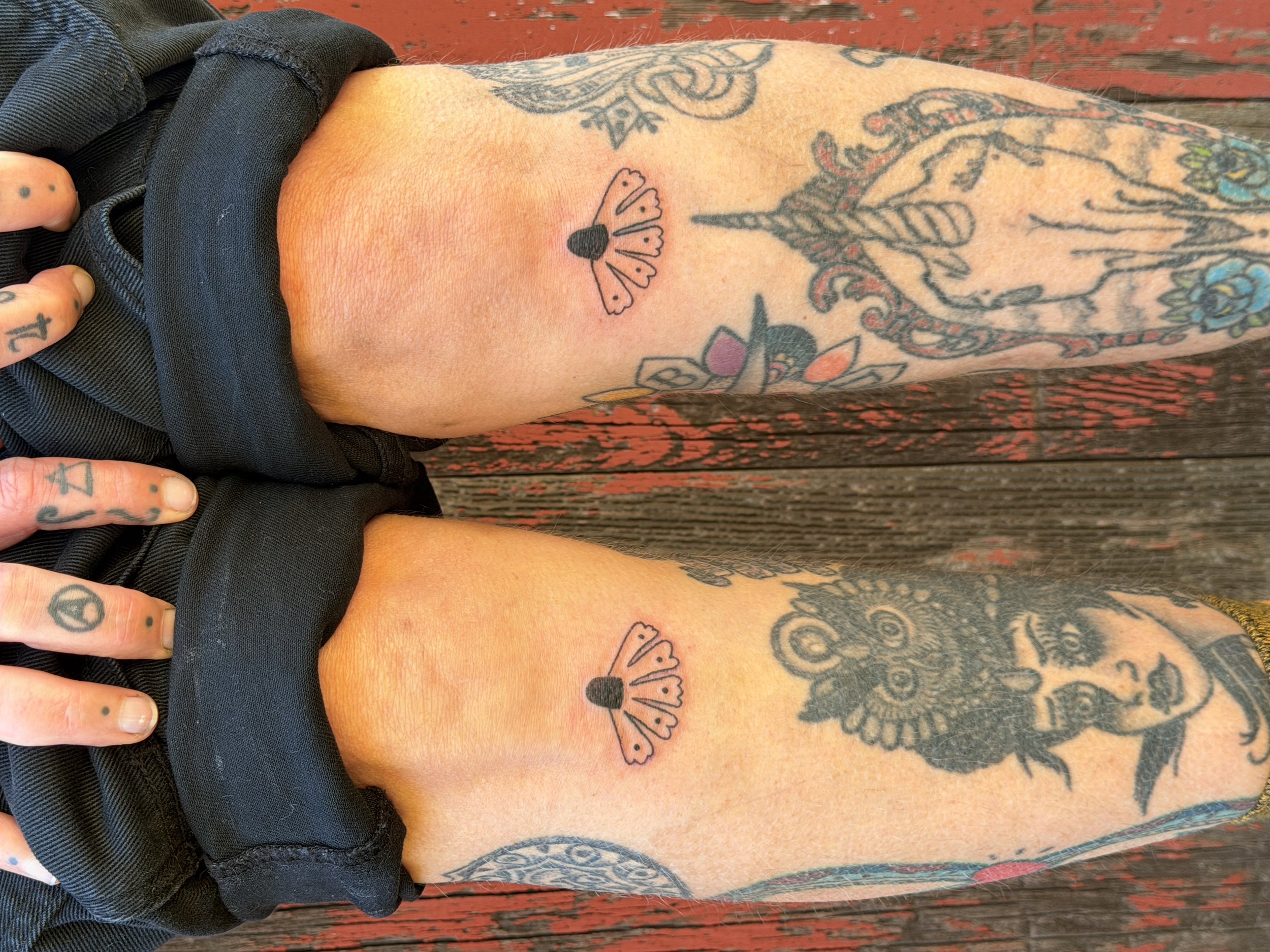 Close-up of a person's arm with multiple tattoos, including a butterfly, a unicorn, floral designs, and decorative patterns. The person is wearing a black shirt and is resting their arm on a wooden surface.